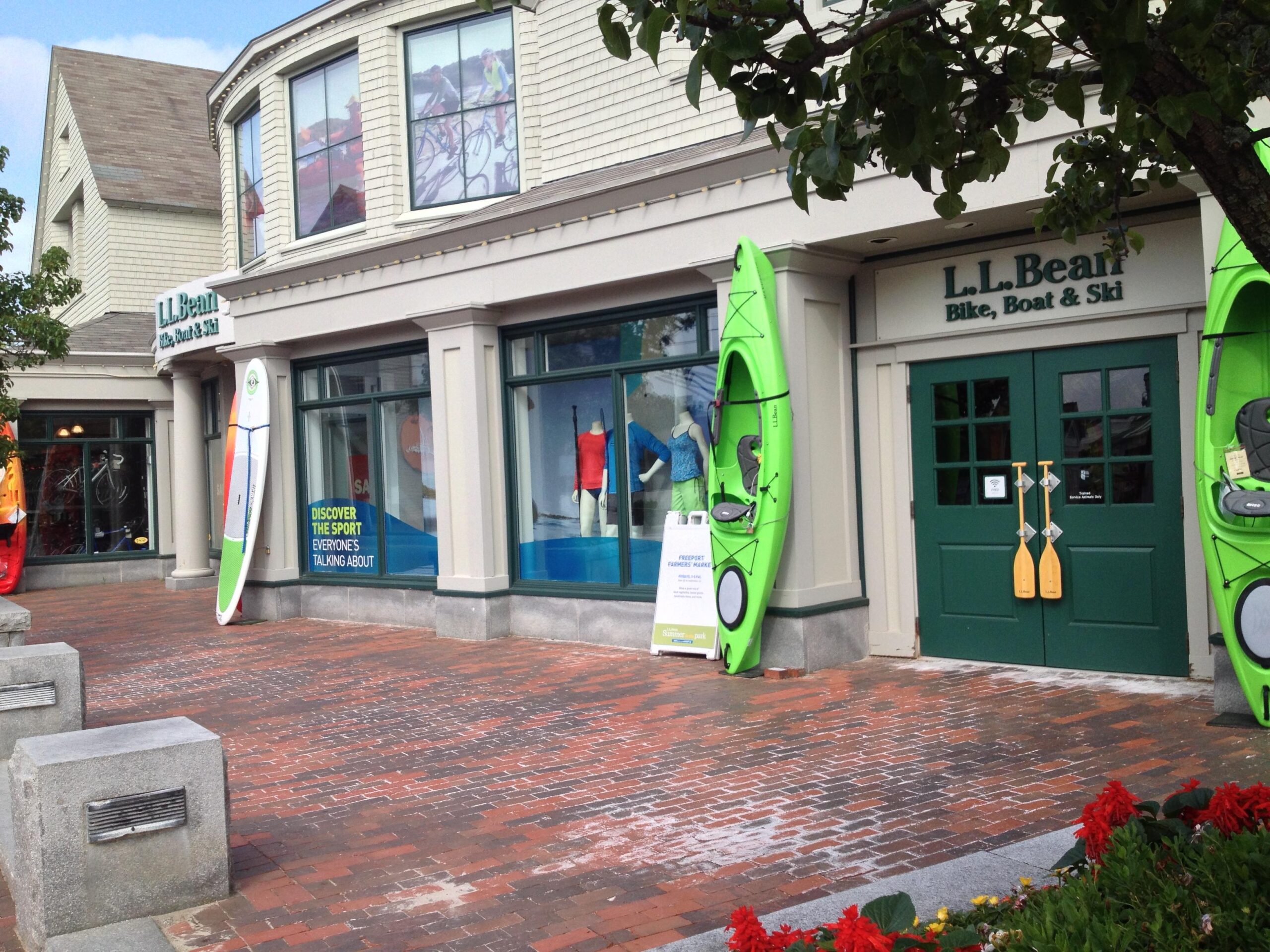 Storefront of an L.L. Bean retail location, featuring two green kayaks displayed at the entrance. The windows showcase colorful outdoor apparel and gear. A sign invites customers to discover a sport everyone is talking about. The area is paved with bricks, and colorful flowers are visible in the foreground, contributing to the outdoor shopping atmosphere.