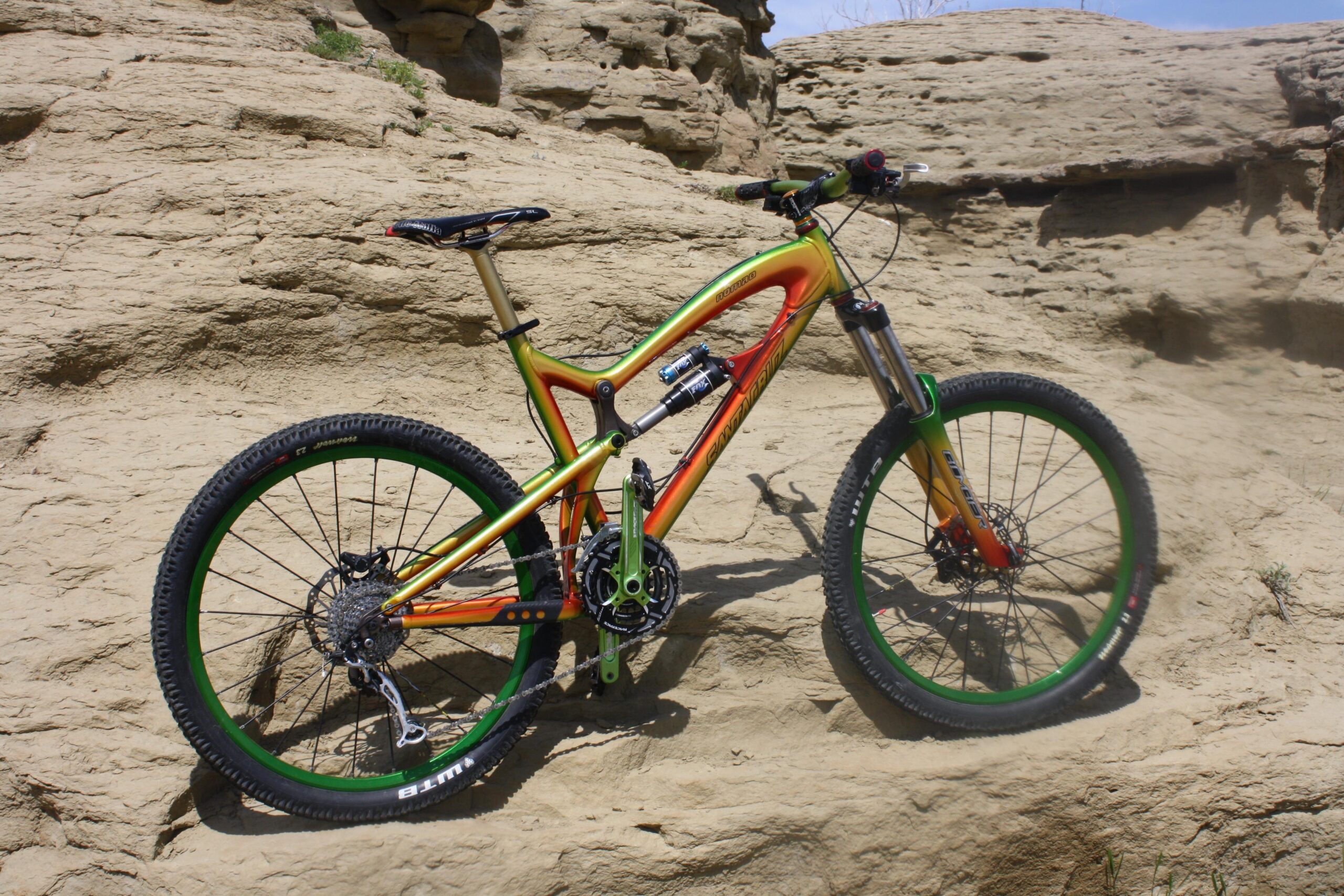 Santa Cruz Nomad: A brightly colored mountain bike with a distinctive frame design, featuring orange, green, and yellow accents, parked on rocky terrain. The bike is positioned on a sandy surface with rugged, uneven features in the background.