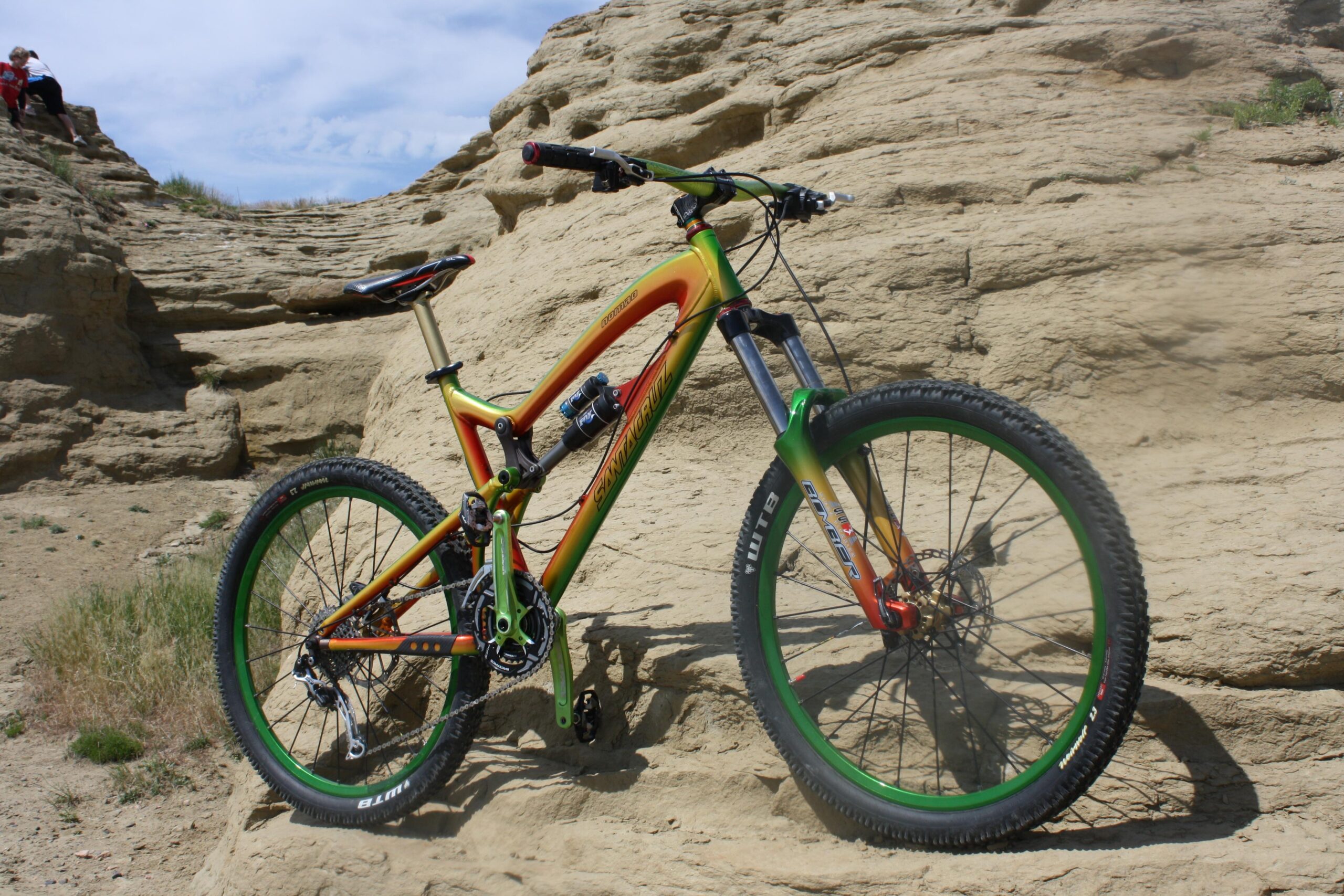 Santa Cruz Nomad: A brightly colored mountain bike resting on rugged, dry terrain with layered rock formations in the background. The bike features a green and orange frame, thick tires, and has a scenic outdoor setting.