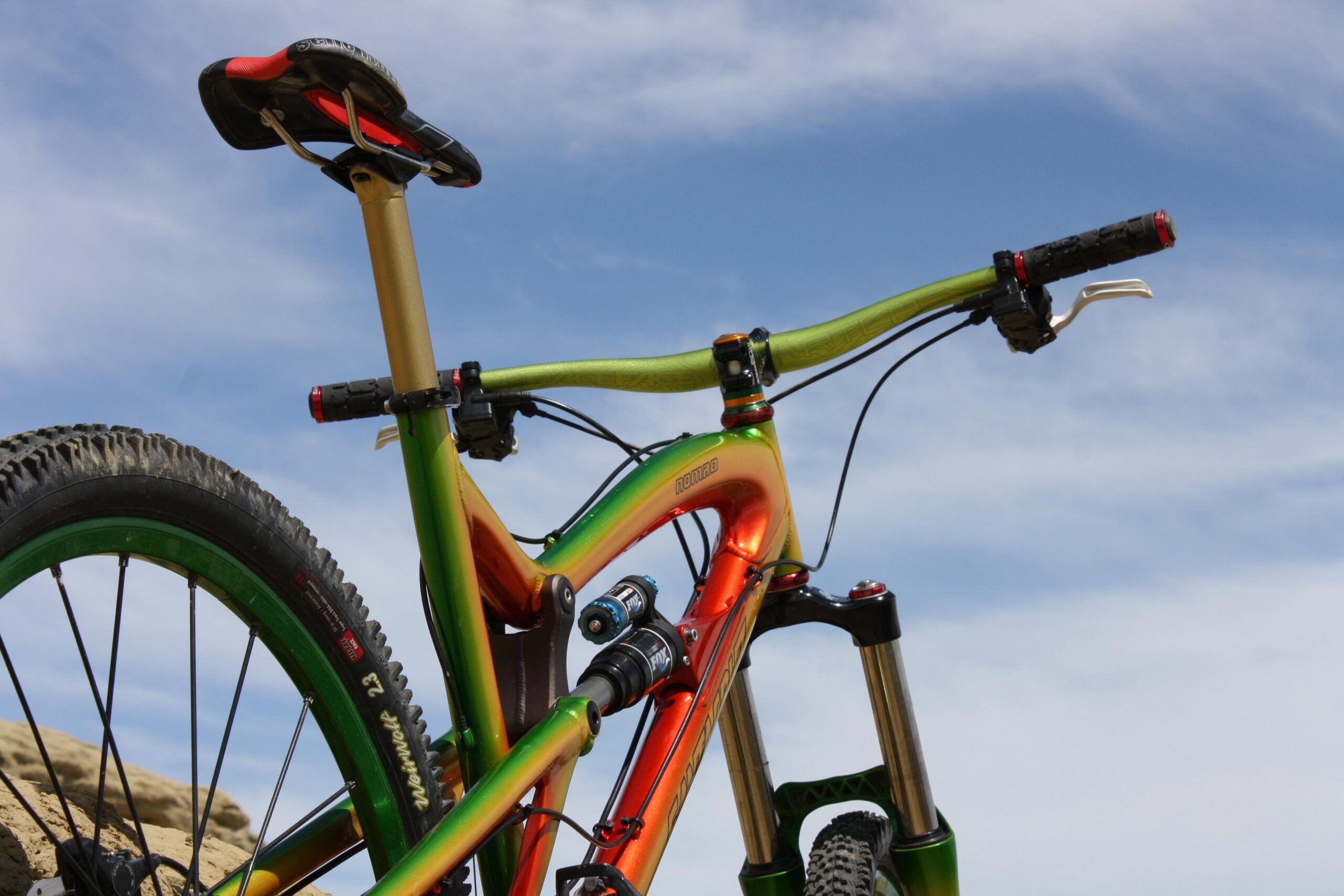 Santa Cruz Nomad: A close-up view of a colorful mountain bike, showcasing its sleek frame in vibrant green and orange hues. The image captures the bike's handlebars, seat, and rear wheel, set against a bright blue sky. The bike features modern components, including a suspension fork and knobby tires, suggesting it's designed for off-road use.