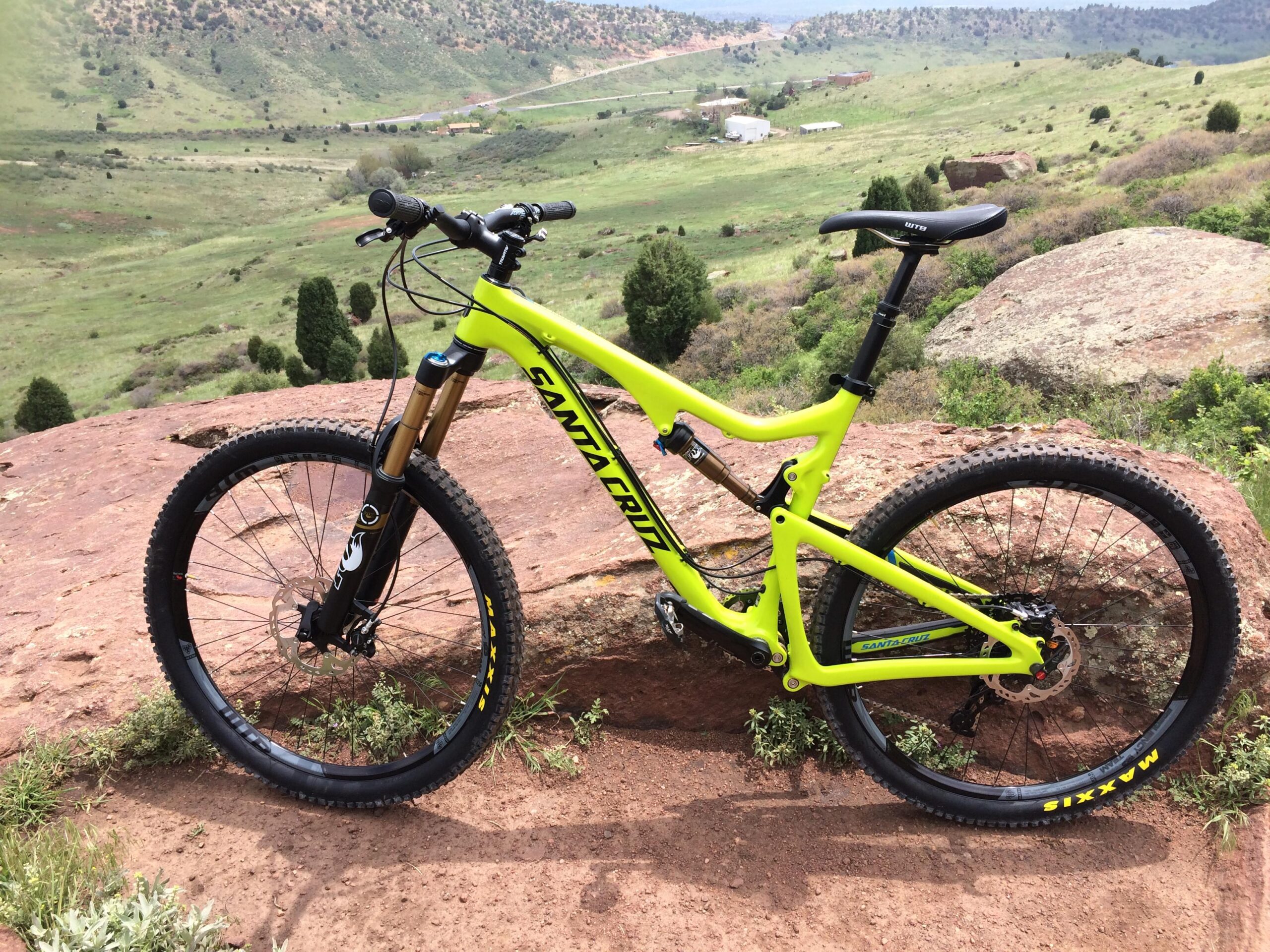 Santa Cruz Bronson: A bright green Santa Cruz mountain bike parked on a rocky outcrop, with a scenic view of rolling hills and sparse trees in the background. The landscape features a mix of grass and rocky terrain under a cloudy sky.