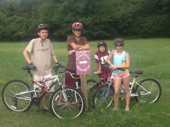 Four children stand with their bicycles in a grassy area, wearing helmets. The eldest boy, dressed in a beige shirt, leans against a wooden sign that points the direction to "Tour d’Wolf." The three others, a boy in a maroon shirt, a girl in a blue top, and a girl in colorful swim shorts, smile as they pose for the photo amidst lush greenery in the background. Tour de Wolf mountain bike trail.