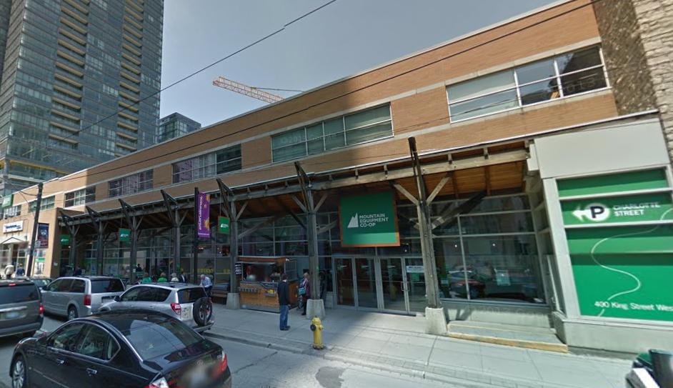 Exterior view of a Mountain Equipment Co-op store, featuring a modern wooden design with large glass windows. The storefront has a prominent green sign displaying the store name. Several cars are parked along the street, and people can be seen entering and exiting the store. Nearby, there is a parking sign for "Charlotte Street." Tall buildings and construction cranes are visible in the background.