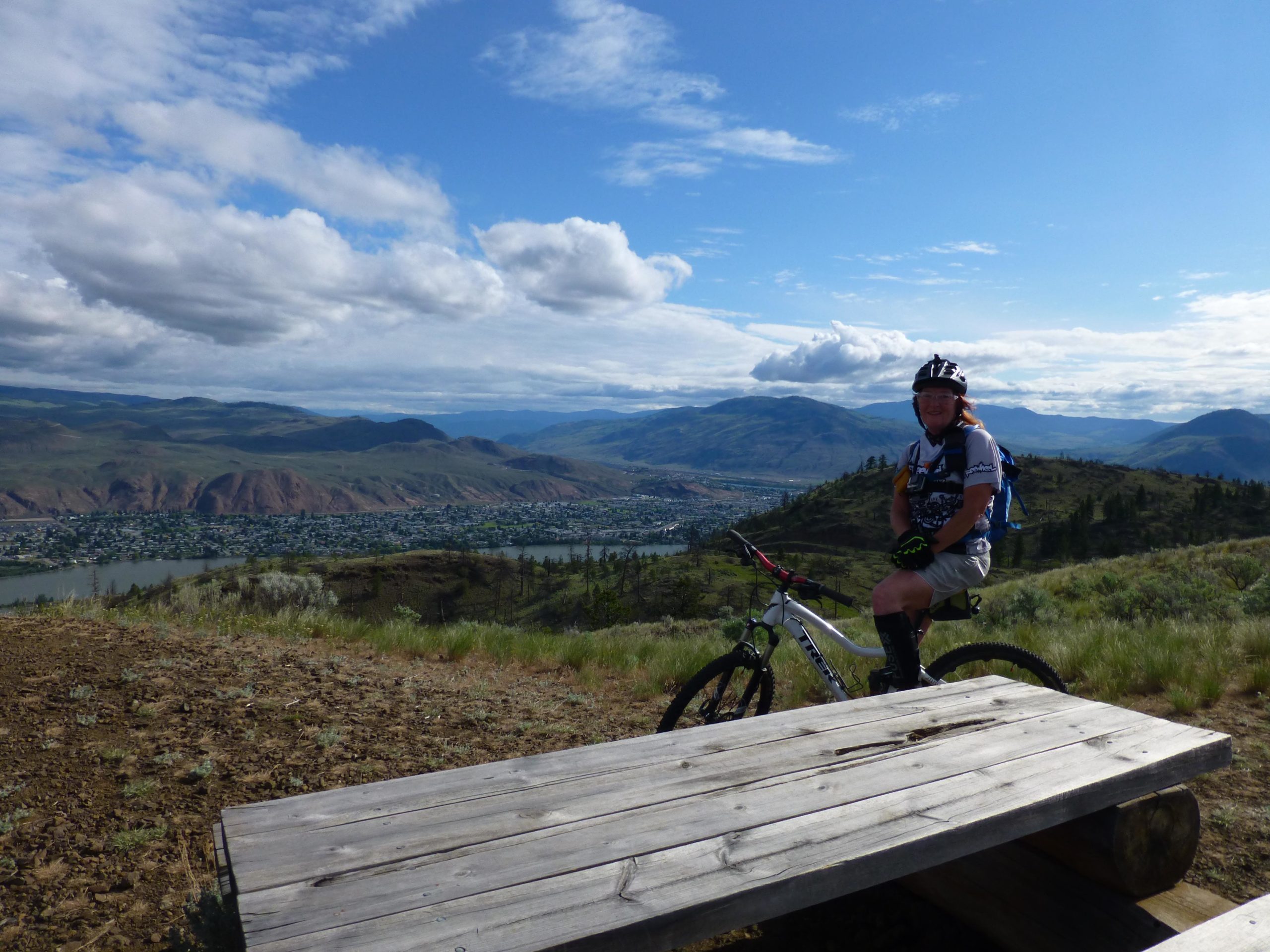 A mountain biker sitting on a bench overlooking a scenic valley. The landscape features rolling hills, a river, and a small town in the distance, all under a partly cloudy sky. The biker is wearing a helmet and cycling gear, with a mountain bike beside them. Kenna Cartwright Park mountain bike trail.