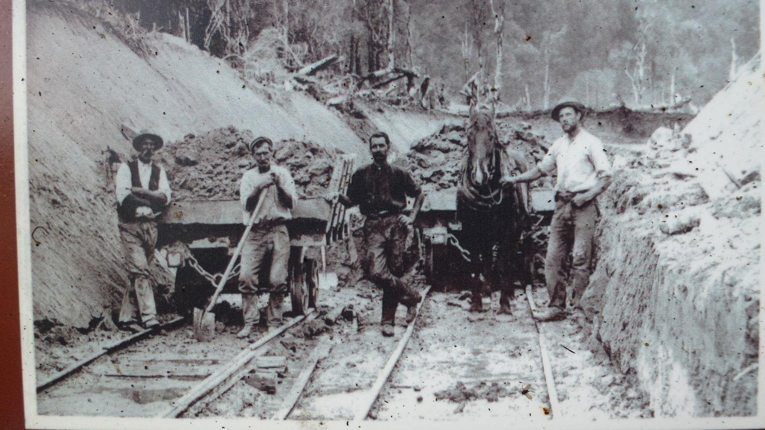 Black and white historical photograph depicting four men standing beside a mining cart on a dirt path. The men are dressed in period work clothing, with two holding shovels and one standing next to a horse. The background features a steep, excavated area with trees and debris. Old Coach Road mountain bike trail.