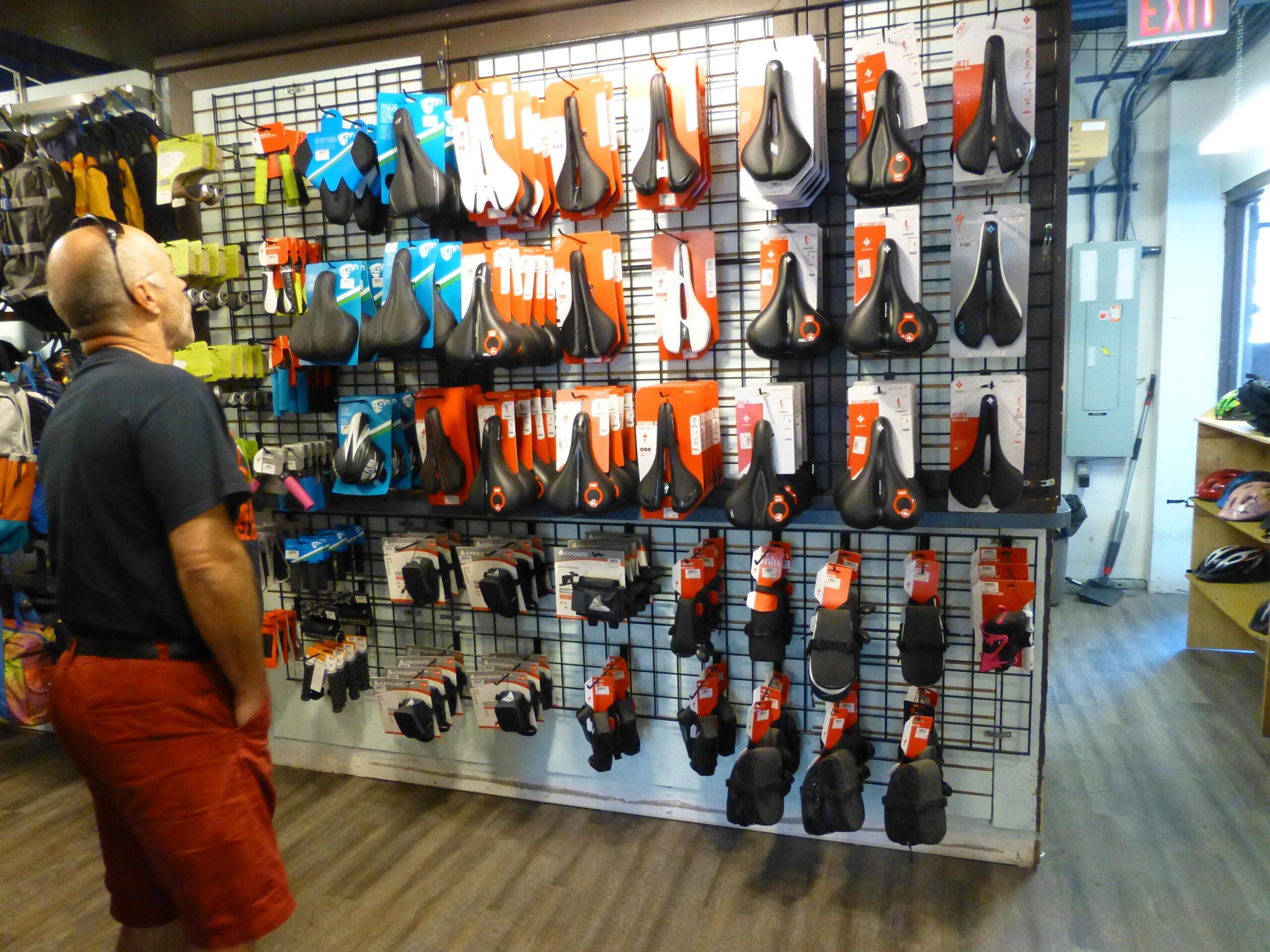 An individual examining a display wall filled with various bicycle seats and accessories in a sporting goods store. The wall features a variety of seat designs and colors, with price tags visible, while shelves in the background hold additional cycling gear.