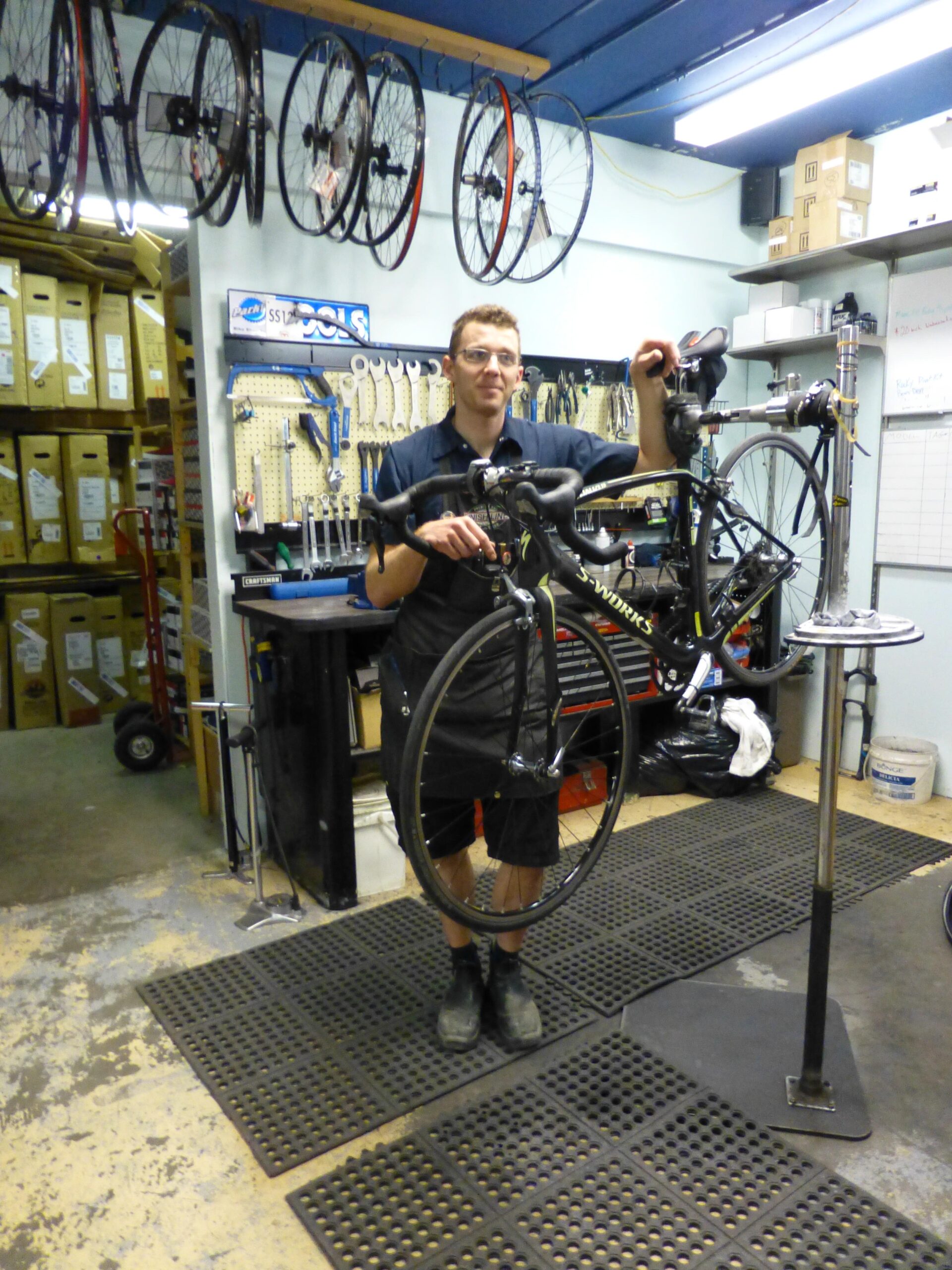 A bicycle mechanic stands in a workshop, holding a bike by its handlebars. The shop is equipped with various bicycle tools and accessories, including hanging wheels and a workbench with organized tools. The mechanic is wearing a black shirt and shorts, and the workspace features rubber matting on the floor.