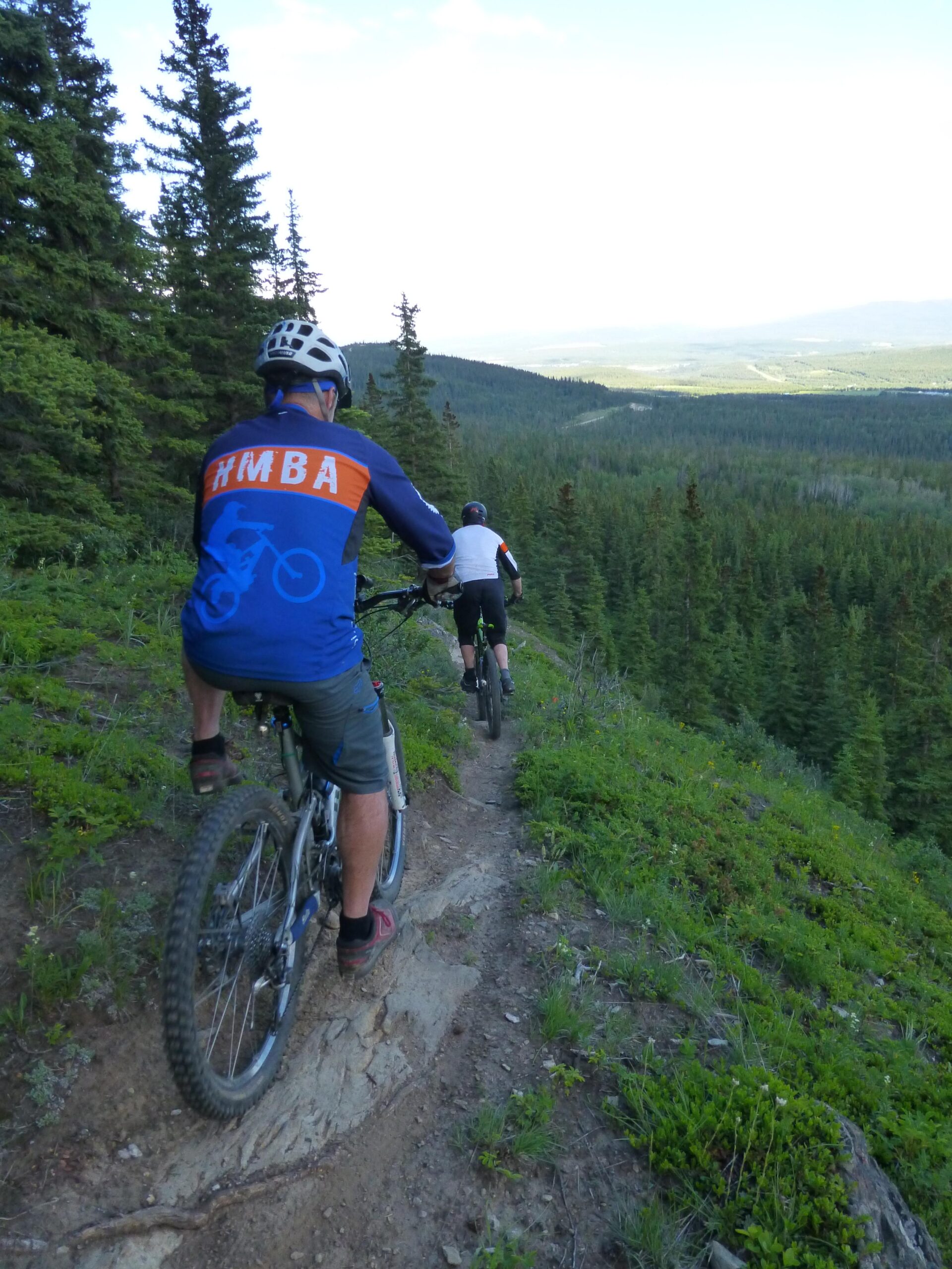 Two mountain bikers descend a narrow, winding trail through a lush green forest, surrounded by tall pine trees. The rider in the foreground is wearing a blue and orange cycling jersey, while the second rider follows closely behind in a white shirt. The scenic landscape stretches out in the background, showcasing rolling hills and valleys under a clear sky. Froehler Coaster mountain bike trail.