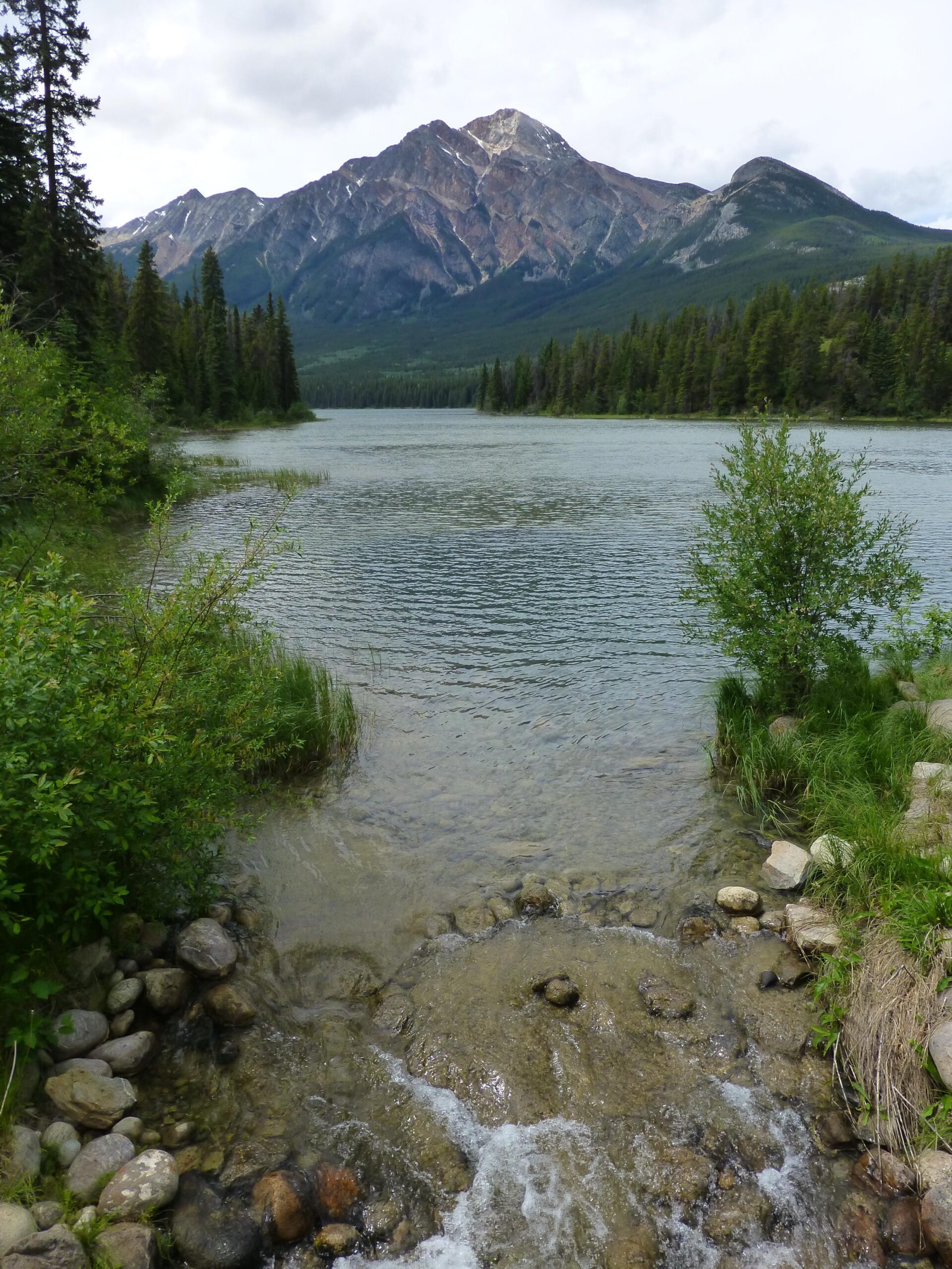 Scenic view of a tranquil lake surrounded by lush greenery, with a gently flowing stream leading into the water. In the background, a majestic mountain range rises under a partly cloudy sky. The landscape showcases a mix of tall trees and rocky terrain, highlighting the natural beauty of the area. Pyramid Bench mountain bike trail.