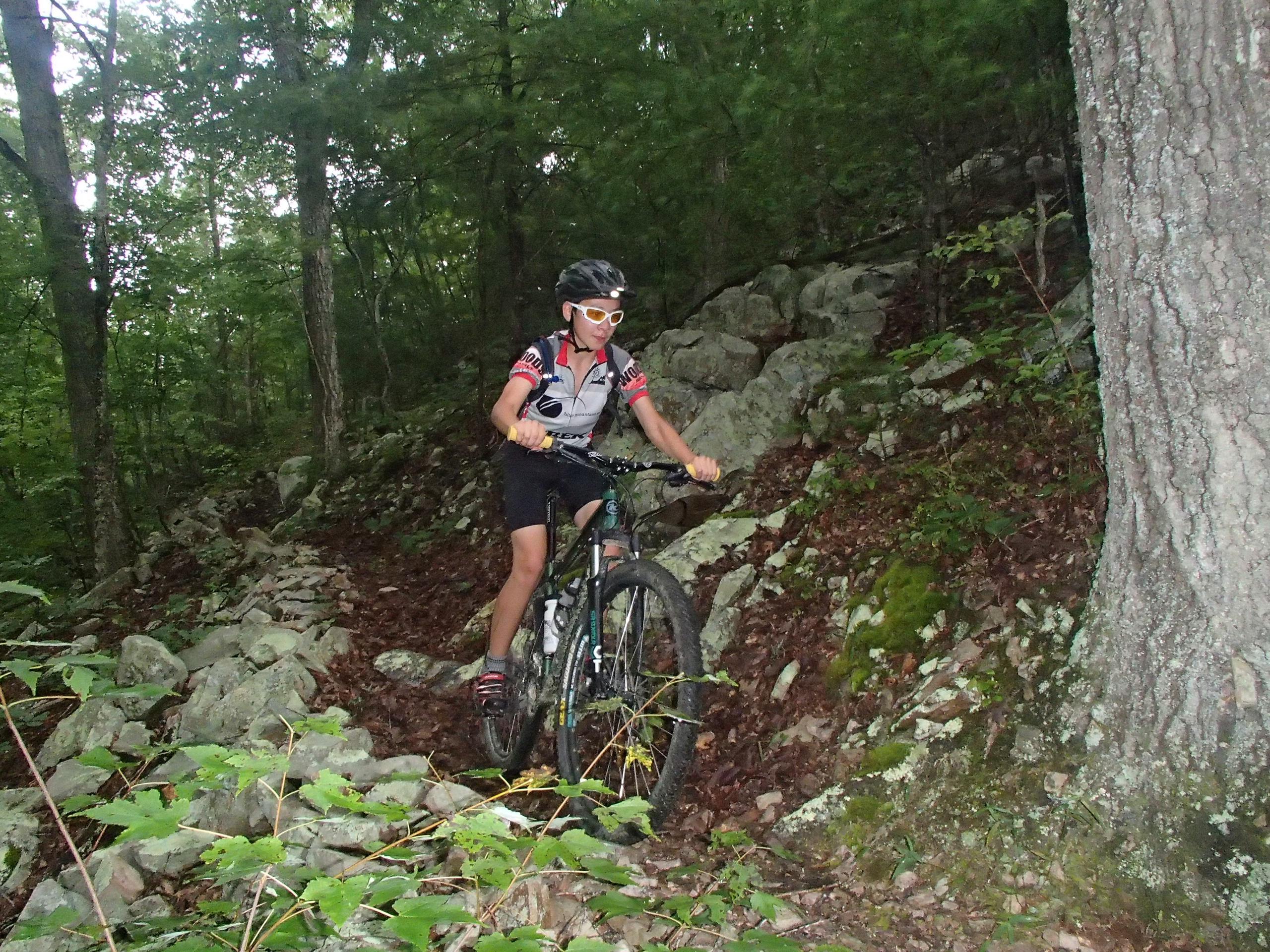 Kona Mahuna: A young cyclist navigating a rocky mountain bike trail surrounded by trees. The rider is wearing a helmet and colorful cycling gear, demonstrating determination and skill as they maneuver through the natural terrain.