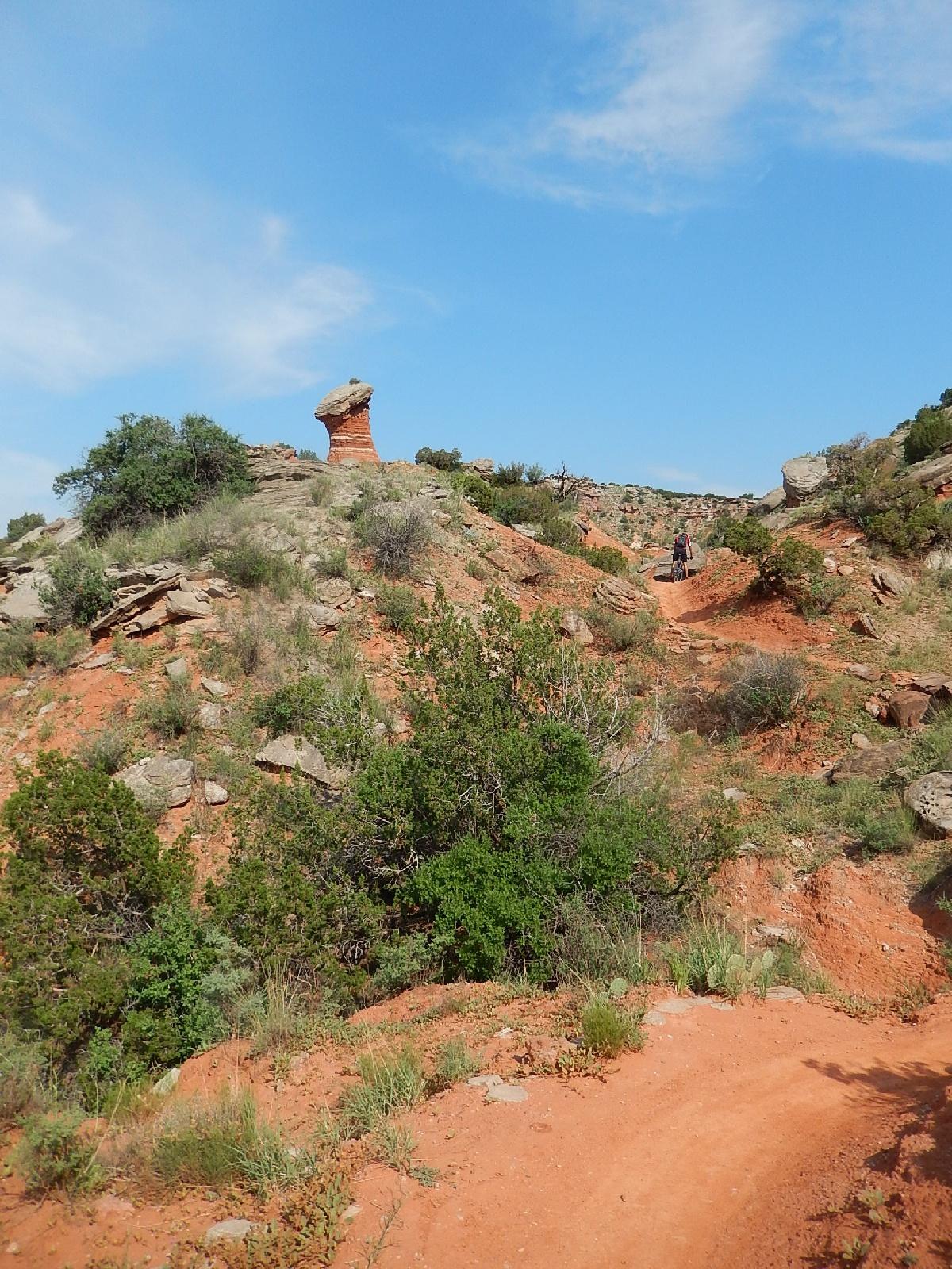A hiking trail winding through a rocky landscape with red soil and sparse greenery, featuring a prominent rock formation resembling a pedestal or mushroom in the background, under a blue sky with wispy clouds. Palo Duro Canyon mountain bike trail.