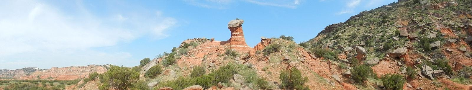 A panoramic view of a unique rock formation resembling a mushroom, situated among red rock cliffs and green shrubs under a blue sky. The landscape features layers of sedimentary rock, highlighting natural geological features. Palo Duro Canyon mountain bike trail.