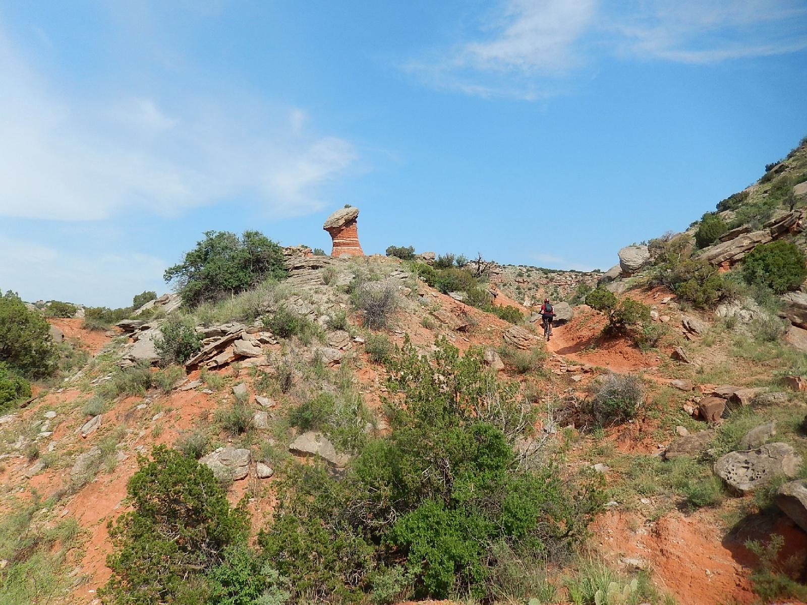 A scenic view of a hilly landscape with reddish soil and rock formations, featuring a prominent rock formation resembling a pedestal. In the distance, a person is walking or biking along a trail that winds through the natural terrain, surrounded by greenery and blue sky. Palo Duro Canyon mountain bike trail.