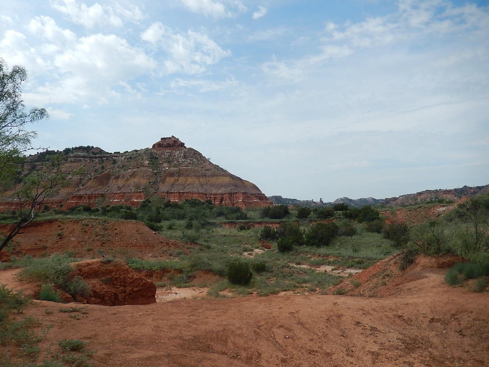 A scenic view of a rugged landscape featuring layered reddish rock formations and green vegetation, with a partly cloudy sky above. The foreground shows a sandy area with sparse bushes, while the background includes a prominent hill with several layers of color and texture. Palo Duro Canyon mountain bike trail.