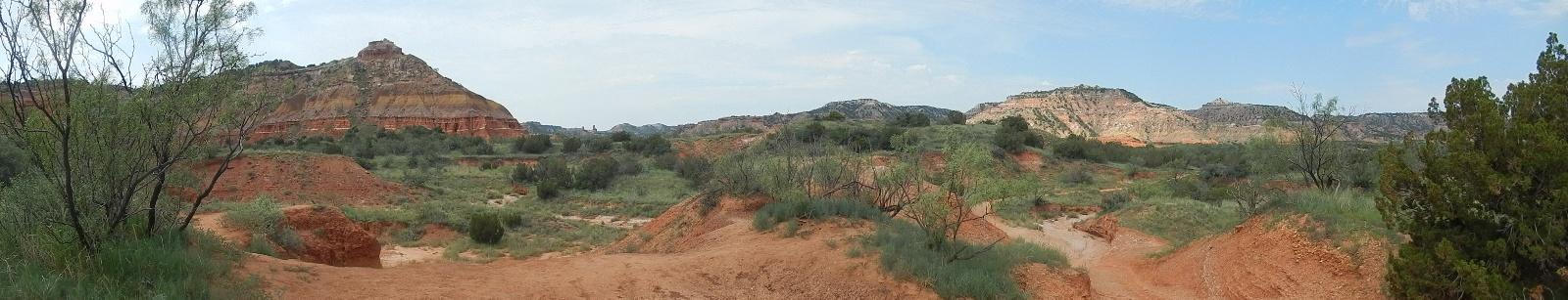 A panoramic view of a rugged, colorful landscape featuring layered rock formations, green vegetation, and a sandy path. The scene showcases a mix of hills and valleys under a partly cloudy sky. Palo Duro Canyon mountain bike trail.