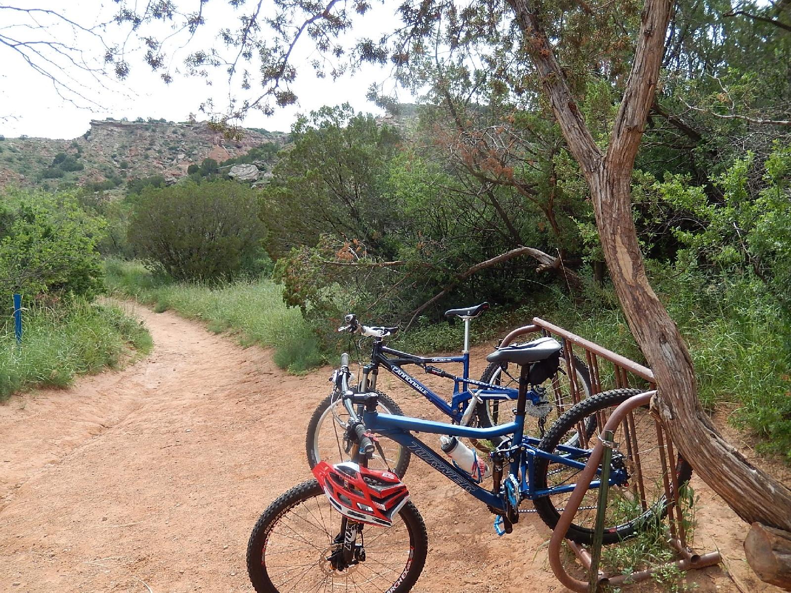 Two bicycles are parked at a bike rack along a dirt path surrounded by greenery and shrubs. A tree is visible to the right, and a rocky hillside can be seen in the background under a cloudy sky. Palo Duro Canyon mountain bike trail.