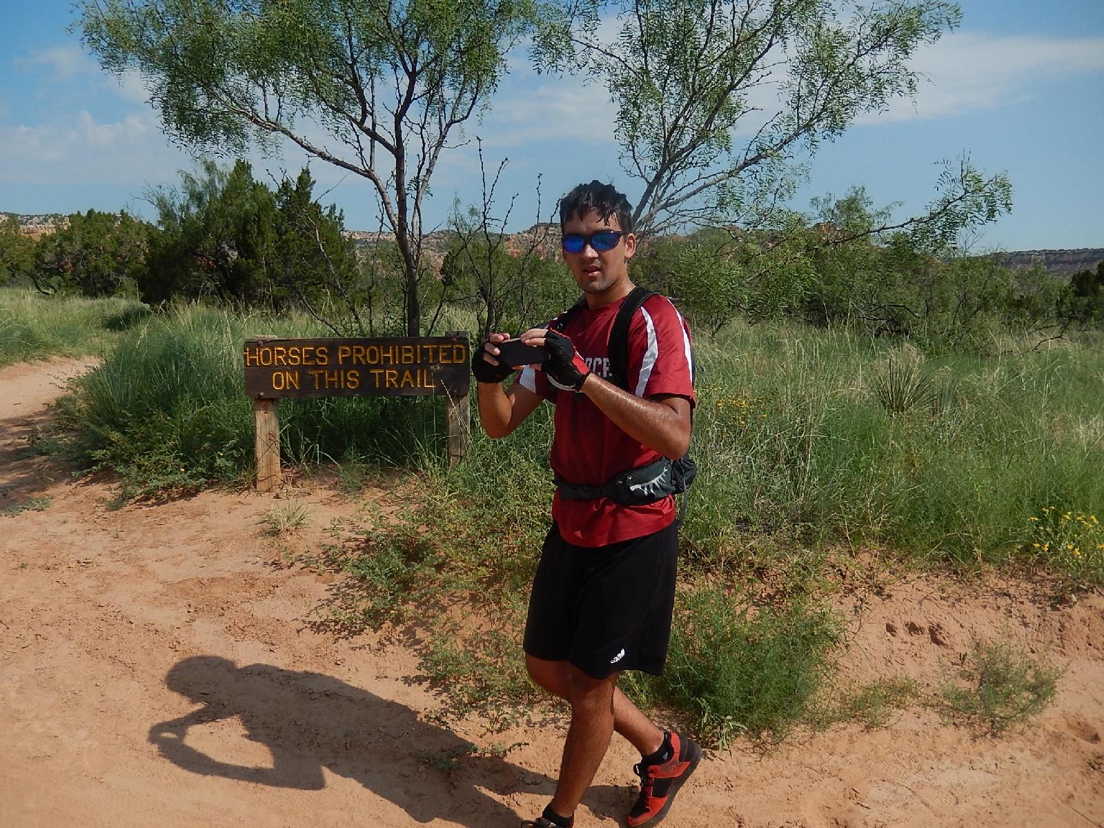 A person in athletic clothing stands on a dirt trail, holding a camera and looking at the viewer. In the background, a sign reads "Horses Prohibited on This Trail," with green vegetation and trees surrounding the area. The sky is partly cloudy, indicating a sunny day. Palo Duro Canyon mountain bike trail.