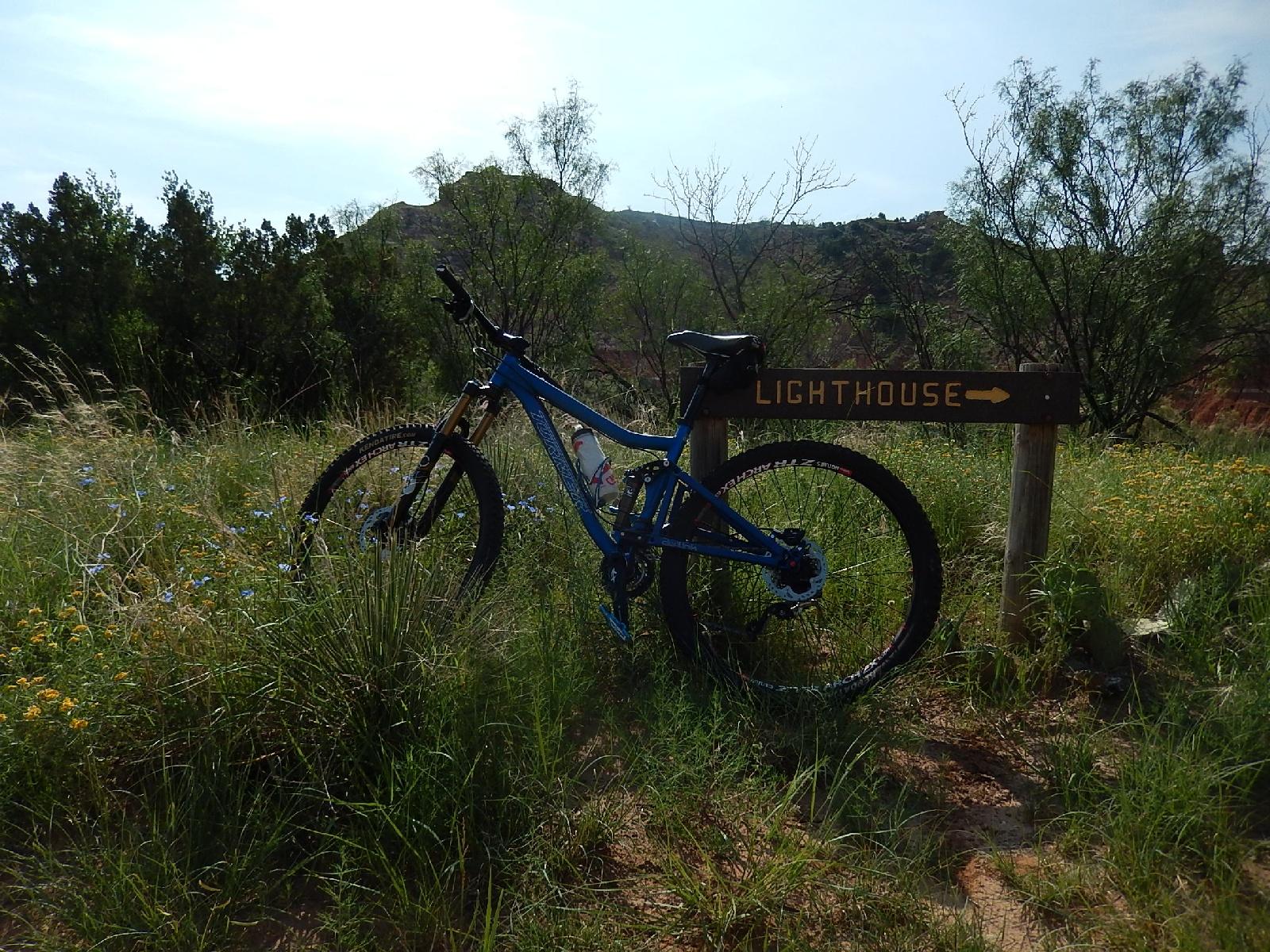 A blue mountain bike parked beside a directional sign that reads "LIGHTHOUSE" pointing to the right, surrounded by tall grass and wildflowers in a natural landscape. Hills can be seen in the background under a clear sky. Palo Duro Canyon mountain bike trail.