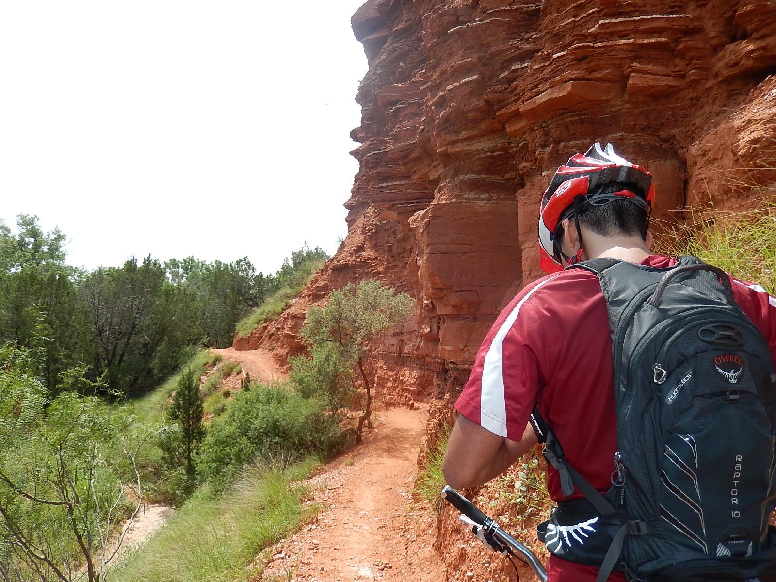 A person wearing a red helmet and a red shirt, standing on a rocky trail surrounded by red rock formations and greenery, with a backpack on their back, preparing to continue biking. Palo Duro Canyon mountain bike trail.
