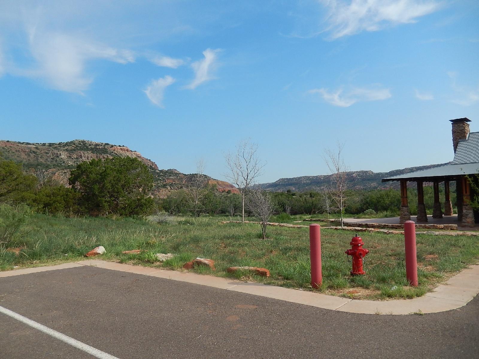 A scenic landscape featuring rolling hills and a blue sky with wispy clouds. In the foreground, there is a paved area with a red fire hydrant and several upright red posts. To the right, a stone pavilion is visible, nestled among patches of greenery and sparse trees. The background showcases rugged hills typical of a natural park setting. Palo Duro Canyon mountain bike trail.