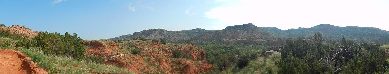 Panoramic view of a rugged landscape featuring red rock formations, green shrubs, and distant hills under a blue sky with light clouds. Palo Duro Canyon mountain bike trail.