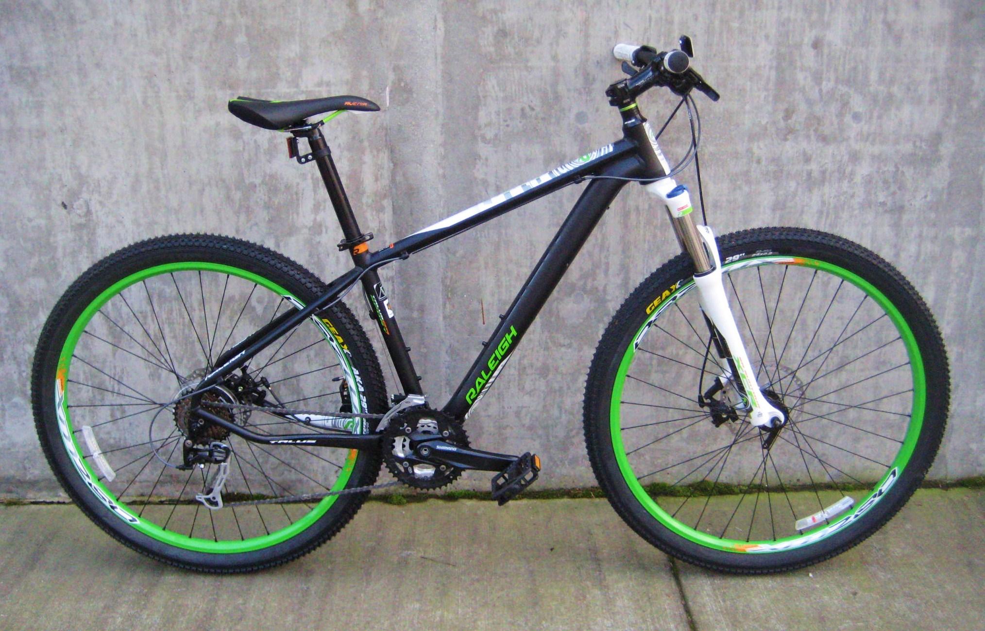 Raleigh Talus: A black mountain bike with green and white accents is positioned against a concrete wall. The bike features thick tires, a comfortable seat, and a front suspension fork, suggesting it's built for off-road use.
