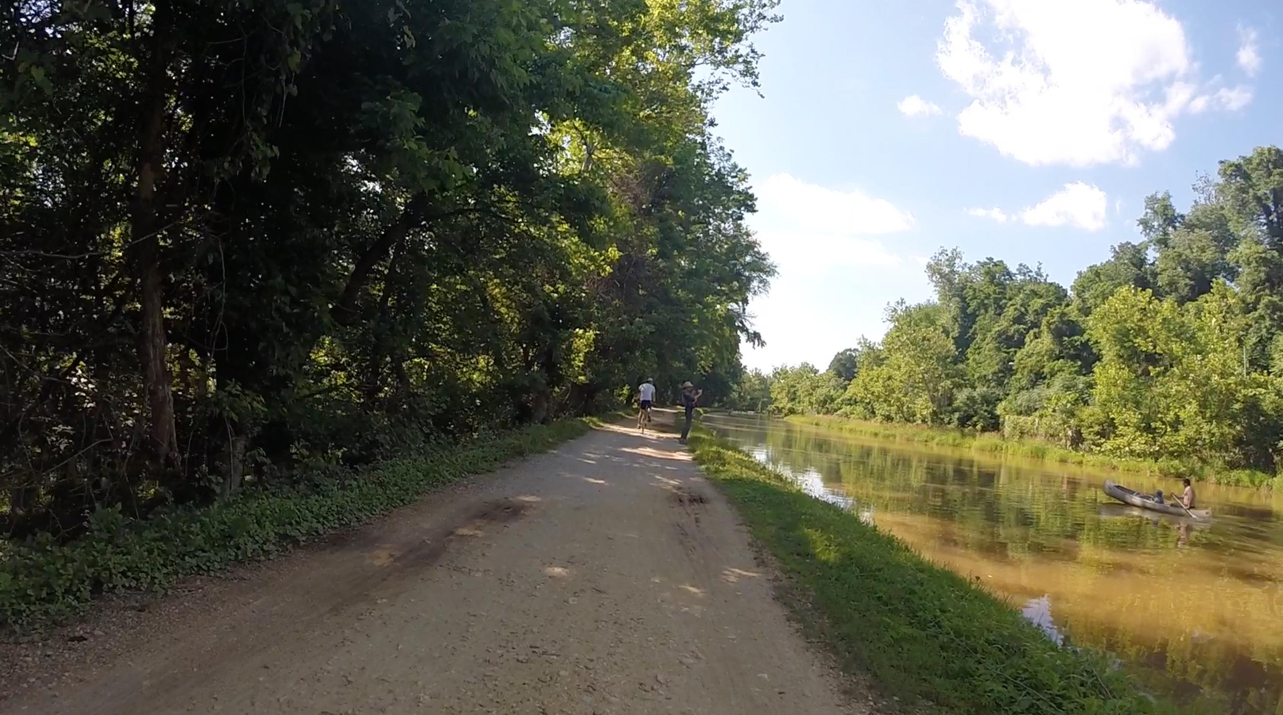 A scenic dirt path lined with trees runs alongside a calm body of water. A cyclist rides on the trail while a person stands nearby. In the water, a canoeist paddles peacefully. The scene is set against a clear blue sky with a few scattered clouds. C&O Canal mountain bike trail.