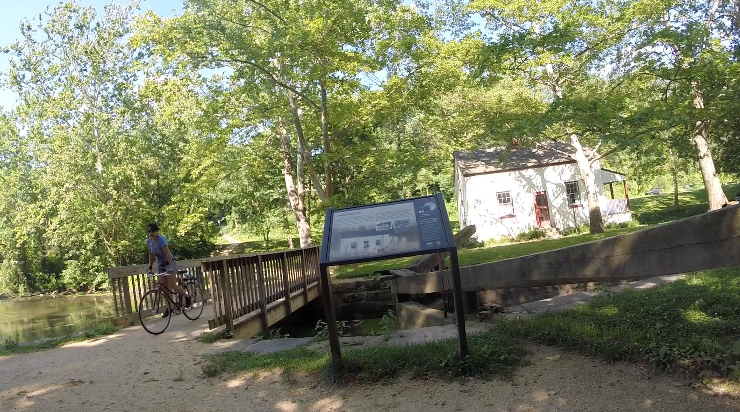 A person riding a bicycle crosses a wooden bridge in a lush, green park. In the foreground, a sign provides information about the area, with a small white building visible in the background surrounded by trees. The scene is bright and sunny, depicting a serene outdoor setting. C&O Canal mountain bike trail.