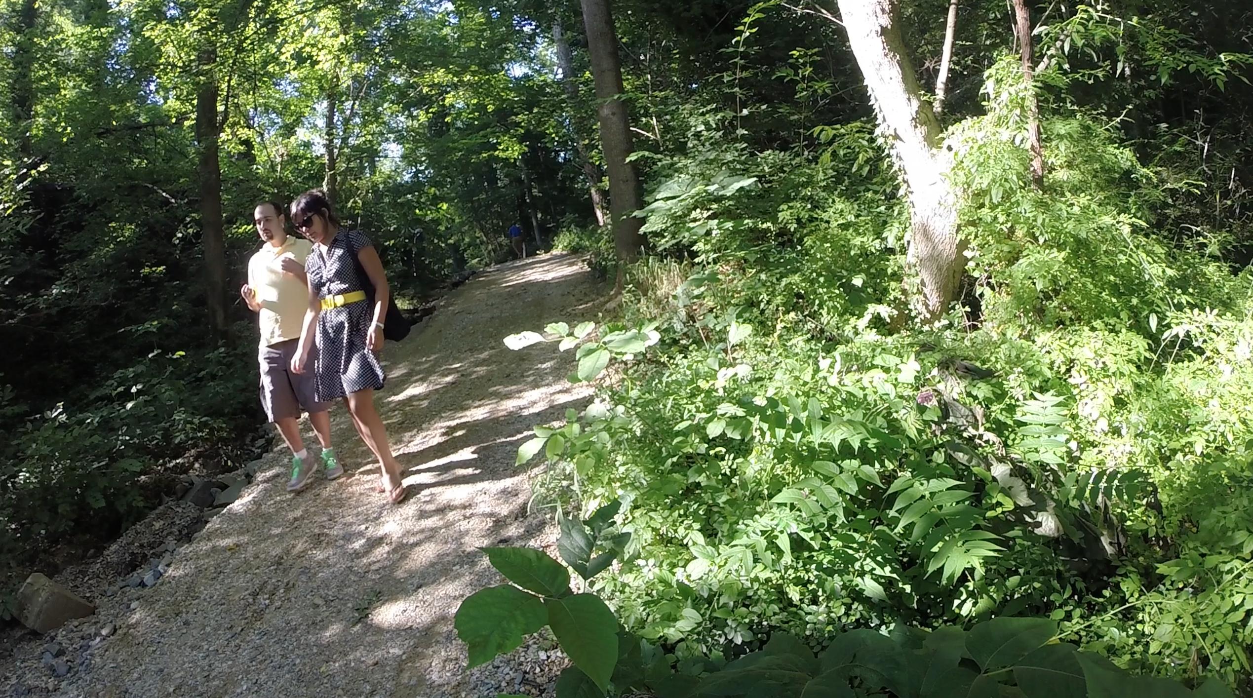 Two people walk along a sunlit gravel path in a lush green forest. They are surrounded by dense foliage and tall trees, with a mix of sunlight and shadows creating a serene atmosphere. One person is wearing a light yellow shirt and shorts, while the other is dressed in a polka dot dress with a bright yellow belt. C&O Canal mountain bike trail.