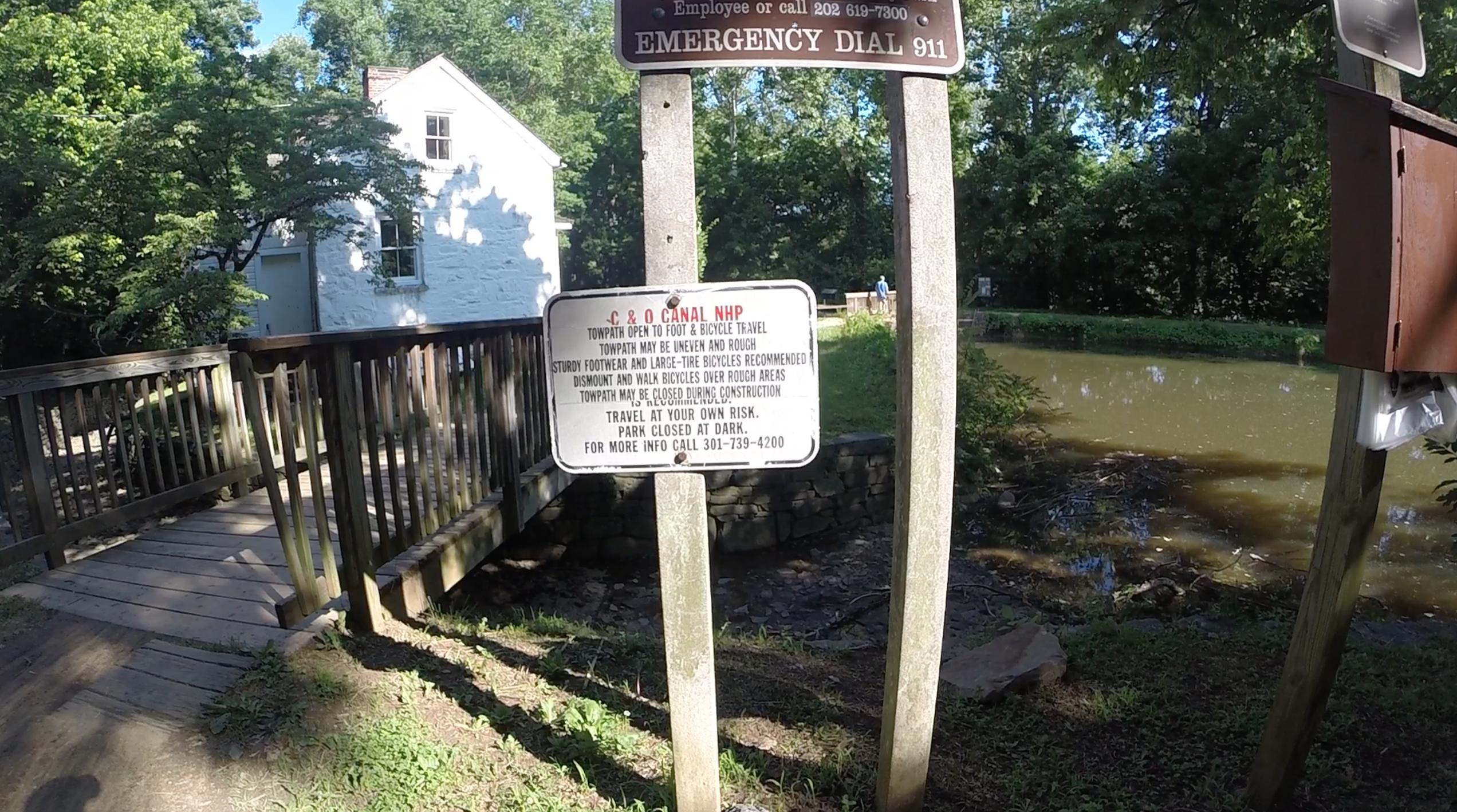 A wooden bridge leading to a towpath, with signs providing safety information for foot and bicycle travel. The signs indicate that the path may be uneven and recommend sturdy footwear. A nearby pond reflects greenery from surrounding trees, and a historic white building is visible in the background. C&O Canal mountain bike trail.