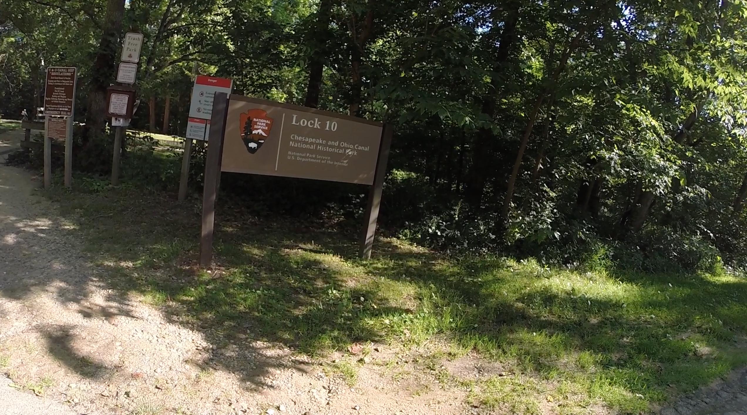 Sign marking Lock 10 of the Chesapeake and Ohio Canal National Historical Park, surrounded by trees and greenery, with a dirt path leading nearby. C&O Canal mountain bike trail.