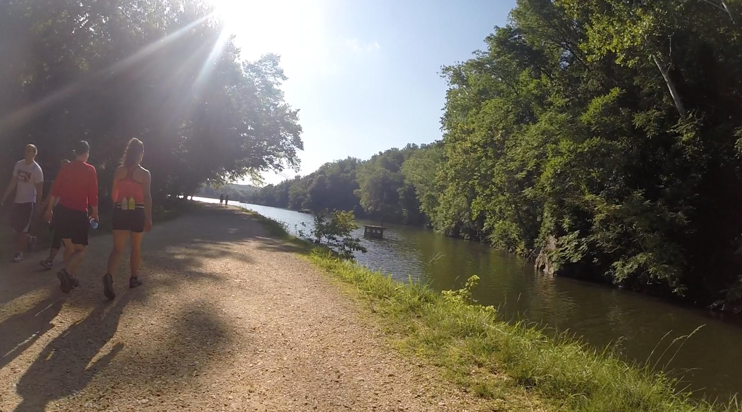 A scenic view of a walking path along a river, bordered by lush green trees. A few people are casually walking on the path, enjoying the sunny day. The sun's rays create a warm glow, adding to the peaceful atmosphere. In the background, a small platform or dock is visible on the water. C&O Canal mountain bike trail.