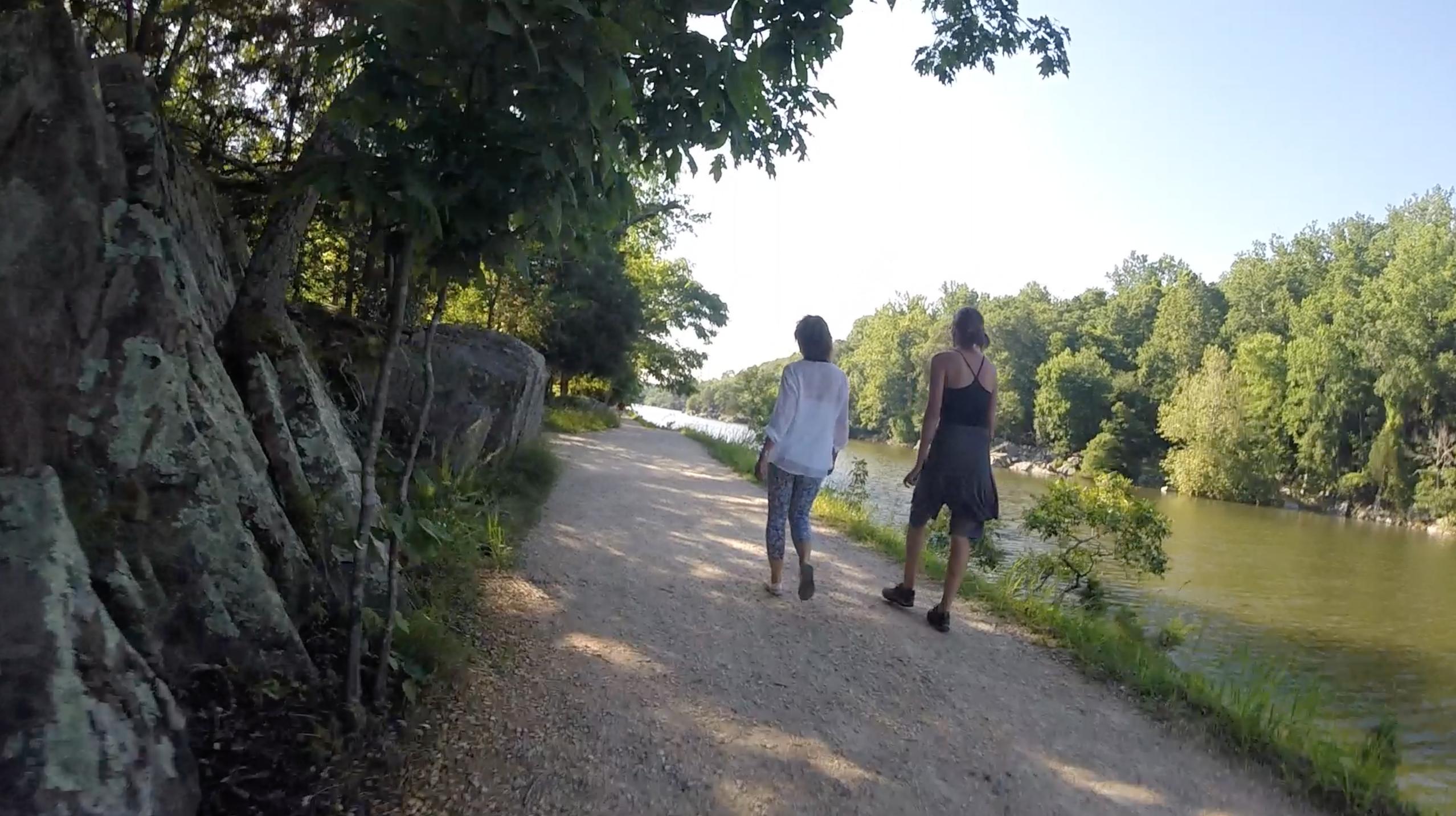 Two individuals walking along a gravel path beside a river, flanked by trees and large rocks. The scene is set in a natural environment with lush greenery under a clear blue sky. C&O Canal mountain bike trail.