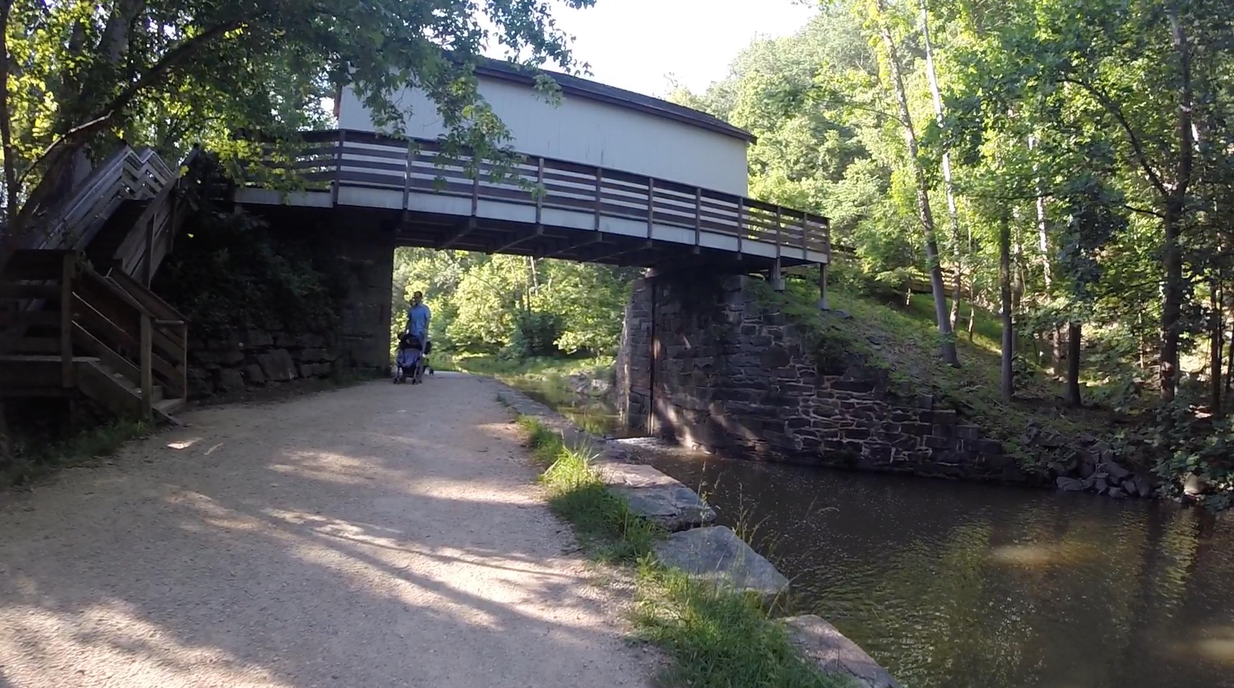 A serene outdoor scene featuring a gravel path beside a calm stream, with a covered bridge overhead. A person pushing a stroller walks along the path, surrounded by lush greenery and trees. The sunlight filters through the leaves, creating a peaceful atmosphere. C&O Canal mountain bike trail.