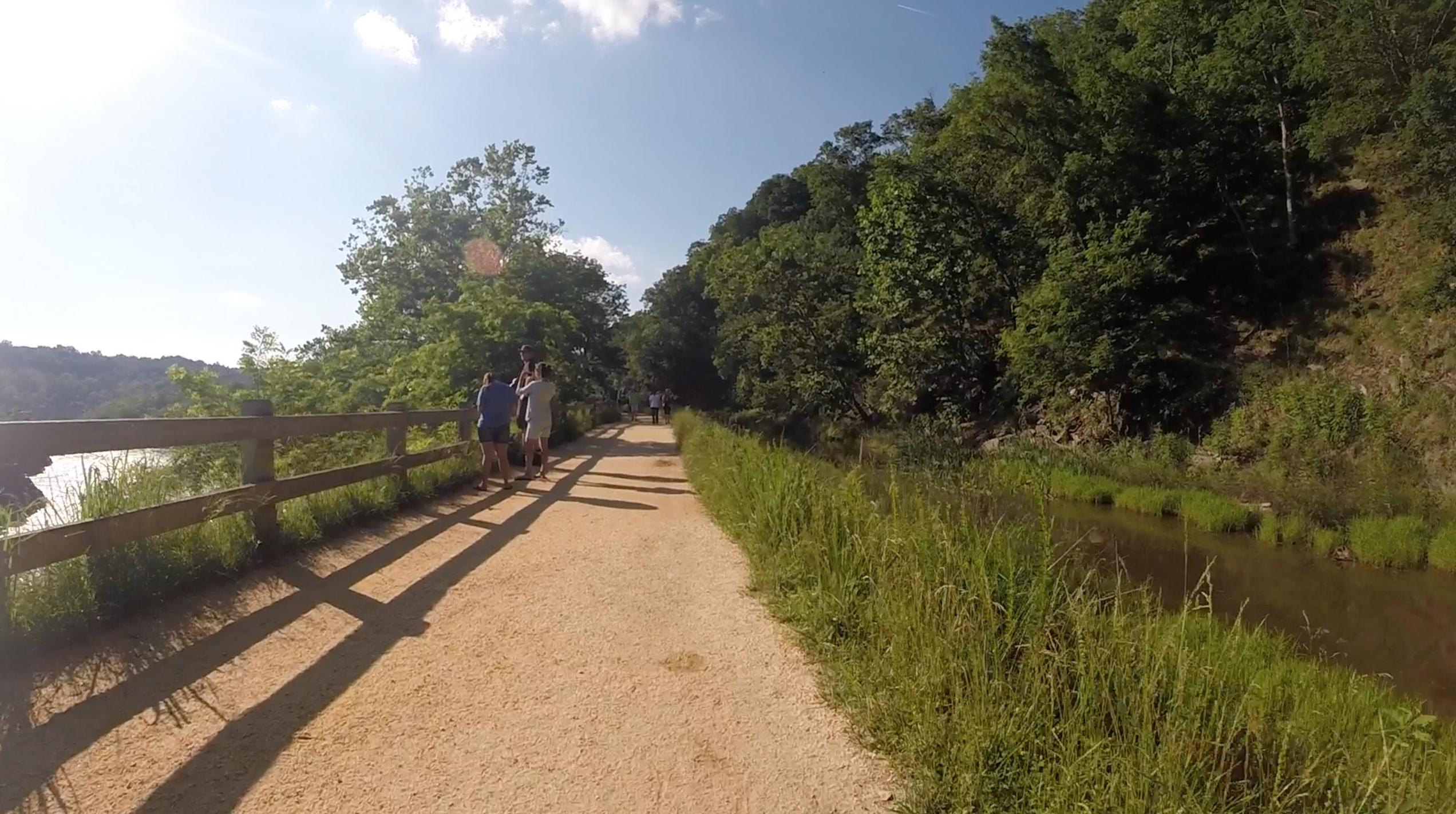A scenic view of a walking path beside a river, surrounded by lush greenery and trees. Two people are standing next to a wooden railing, enjoying the view, while others stroll along the path. The bright sunlight casts long shadows on the ground, creating a warm and inviting atmosphere. C&O Canal mountain bike trail.
