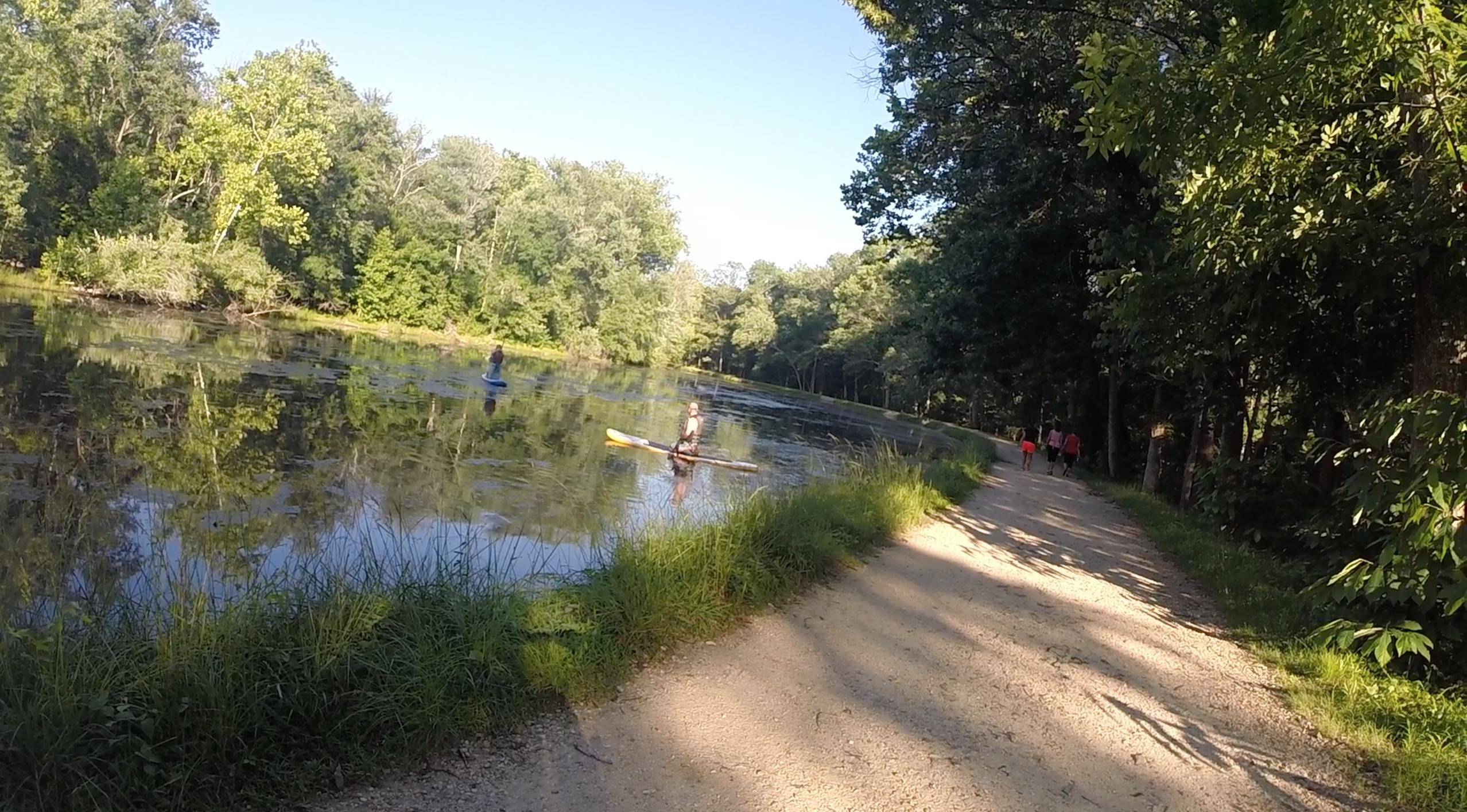 A peaceful scene depicting a calm pond surrounded by lush greenery. Two individuals are paddleboarding on the water, while a walking path bordered by grass runs alongside the pond. The sun shines brightly, reflecting off the water's surface, creating a serene outdoor atmosphere. C&O Canal mountain bike trail.