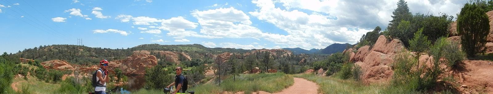 Two cyclists are standing on a dirt path surrounded by lush greenery and red rock formations under a blue sky with fluffy white clouds. The landscape features hills and distant mountains, creating a scenic outdoor environment. Red Rock Canyon mountain bike trail.