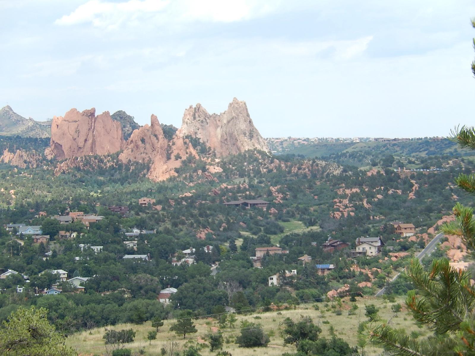 A scenic view of a rocky landscape featuring red rock formations in the background and a small residential area surrounded by greenery in the foreground. The sky is partly cloudy, and the scene captures a blend of natural beauty and suburban life. Red Rock Canyon mountain bike trail.