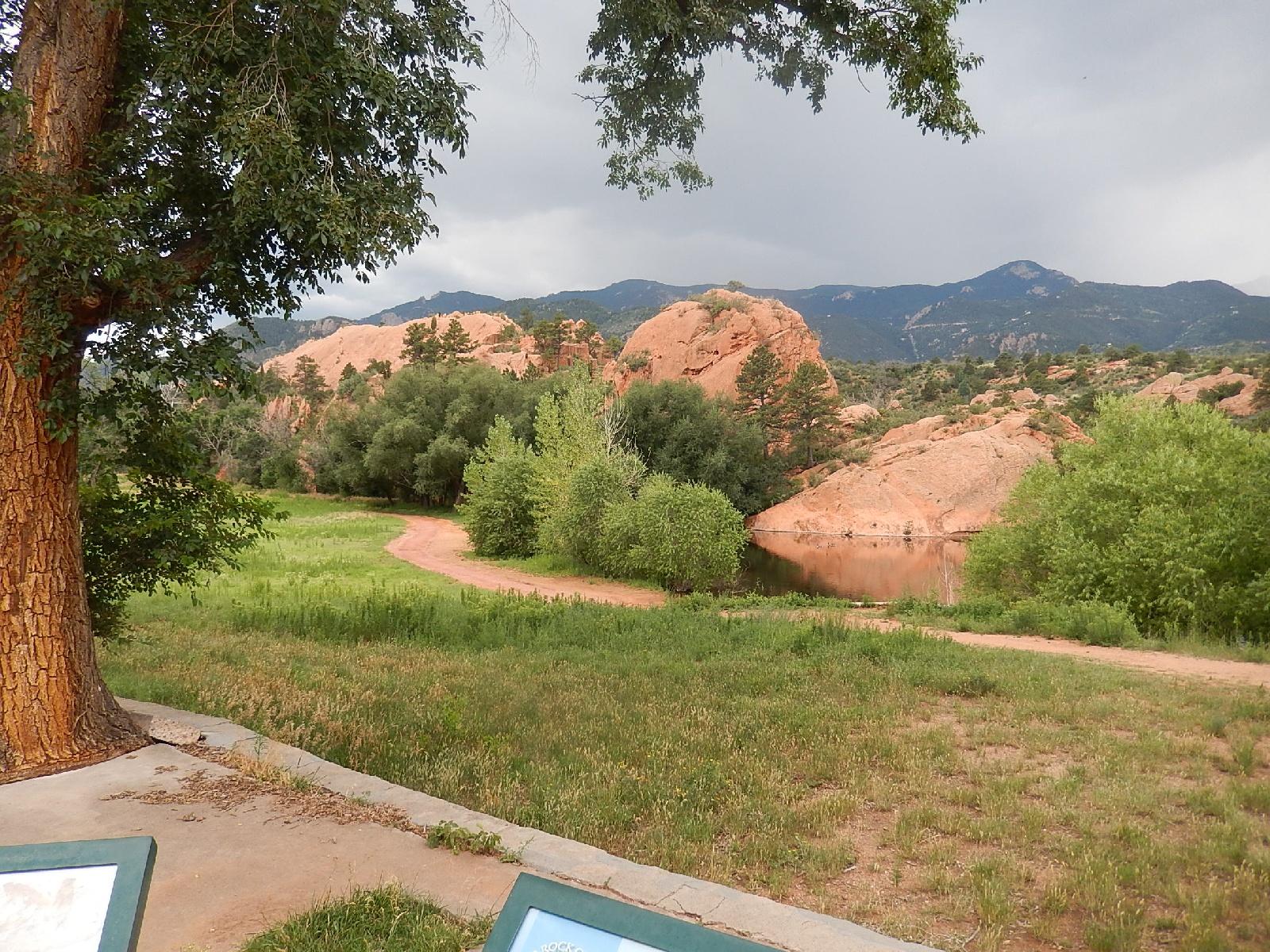 A scenic view of a natural landscape featuring rocky formations in the background, lush greenery in the foreground, and a winding path leading to a calm body of water. Overcast skies hint at the possibility of rain, while nearby trees frame the scene beautifully. Two informational signs are partially visible in the foreground. Red Rock Canyon mountain bike trail.
