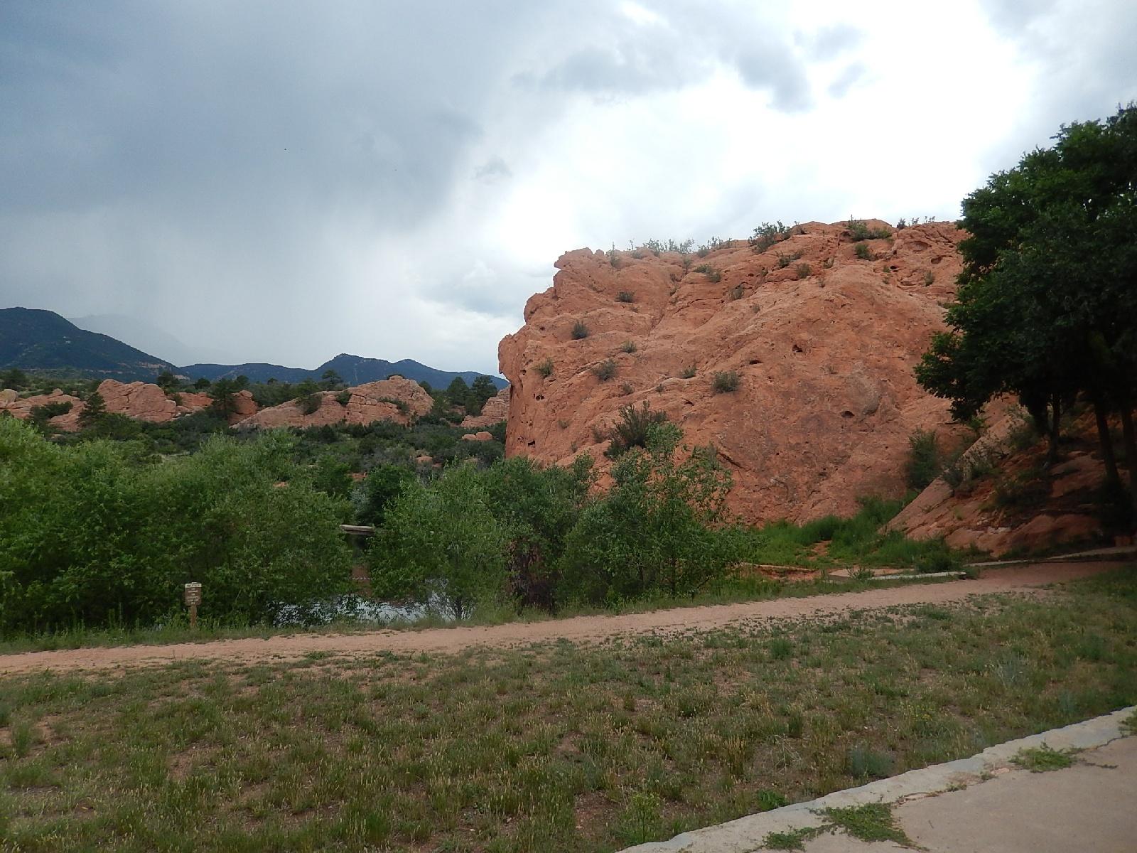 A scenic view of a natural landscape featuring a large red rock formation surrounded by greenery. In the foreground, a path runs alongside lush vegetation, leading to a small body of water. The sky is cloudy, suggesting the possibility of rain, with distant mountains visible in the background. Red Rock Canyon mountain bike trail.