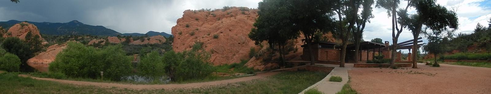 Panoramic view of a natural landscape featuring red rock formations and green foliage. In the foreground, there's a pathway leading to a covered structure nestled among trees, with a serene body of water visible nearby. The background showcases mountains beneath a cloudy sky, hinting at an overcast day. Red Rock Canyon mountain bike trail.