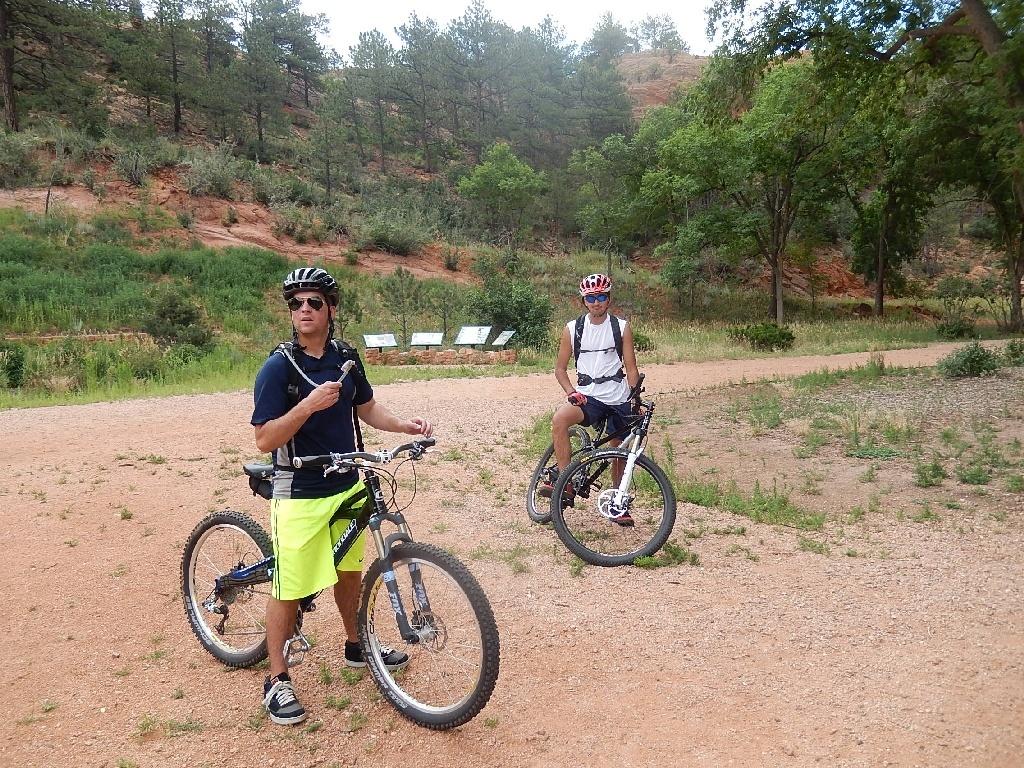 Two cyclists stand on a dirt path in a natural setting, surrounded by greenery and trees. One cyclist, wearing a black helmet and sunglasses, is holding a water bottle and wearing a blue shirt with yellow shorts. The other cyclist, wearing a red helmet and sunglasses, is seated on a white mountain bike in a white shirt and dark shorts. In the background, a rocky hillside can be seen along with some signboards. Red Rock Canyon mountain bike trail.