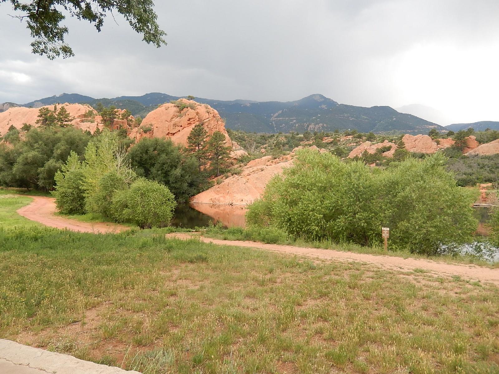 A scenic landscape featuring rocky hills and greenery under a cloudy sky. A dirt path meanders through lush grass and trees, leading to a calm body of water reflecting the surrounding nature. In the distance, mountains rise, creating a picturesque backdrop. Red Rock Canyon mountain bike trail.