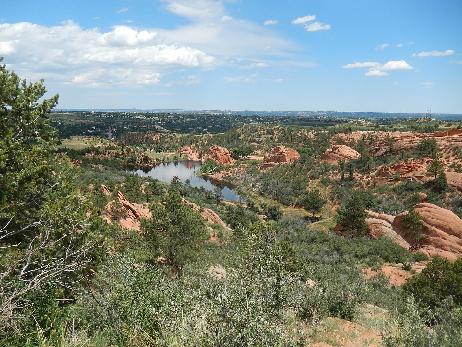 A scenic view of a natural landscape featuring red rock formations, lush greenery, and a tranquil pond under a partly cloudy sky. The image showcases rolling hills and distant mountains, inviting a sense of peace and connection with nature. Red Rock Canyon mountain bike trail.
