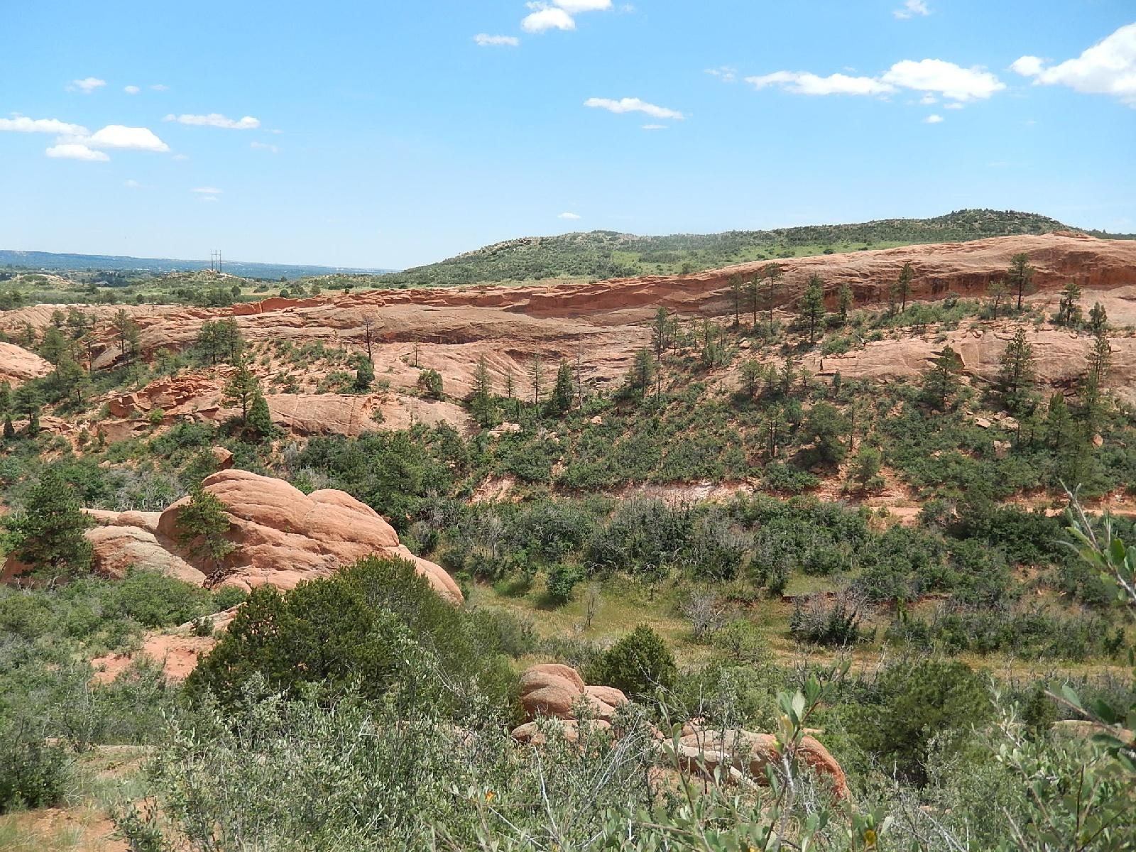 A scenic view of a rocky landscape featuring reddish rock formations, patches of greenery, and pine trees under a bright blue sky with scattered clouds. The terrain includes gently rolling hills in the background. Red Rock Canyon mountain bike trail.