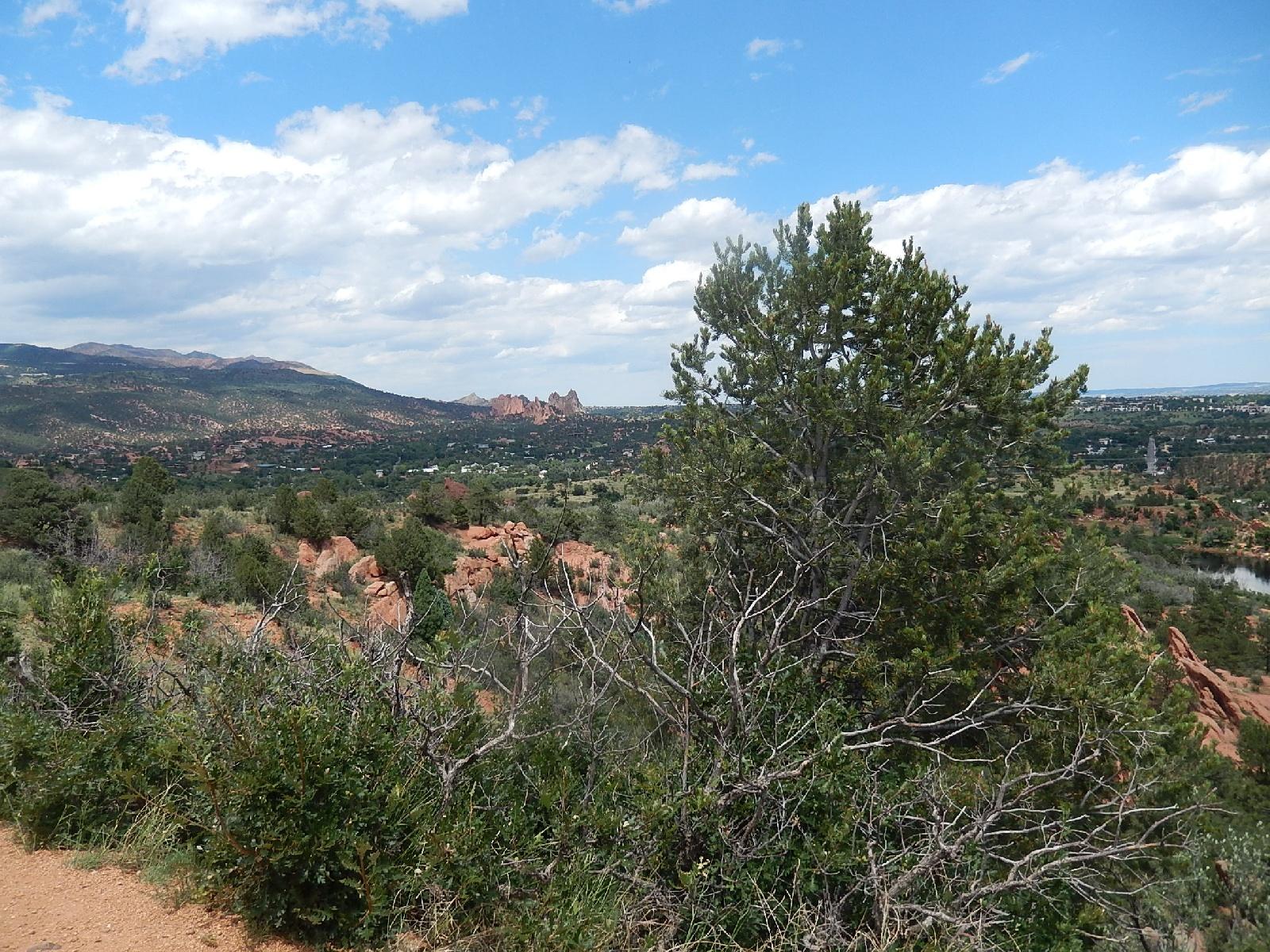 A panoramic view of a mountainous landscape featuring green trees and rocky terrains under a partly cloudy sky. In the distance, red rock formations are visible, with a small body of water meandering through the valley below. The scene captures the natural beauty and tranquility of the outdoors. Red Rock Canyon mountain bike trail.