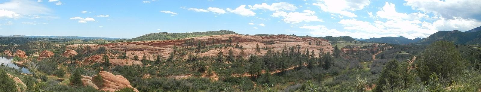 A panoramic view of a rugged landscape featuring red rock formations, green trees, and a winding river. The scene is set under a bright blue sky with scattered clouds, showcasing rolling hills and distant mountains in the background. Red Rock Canyon mountain bike trail.