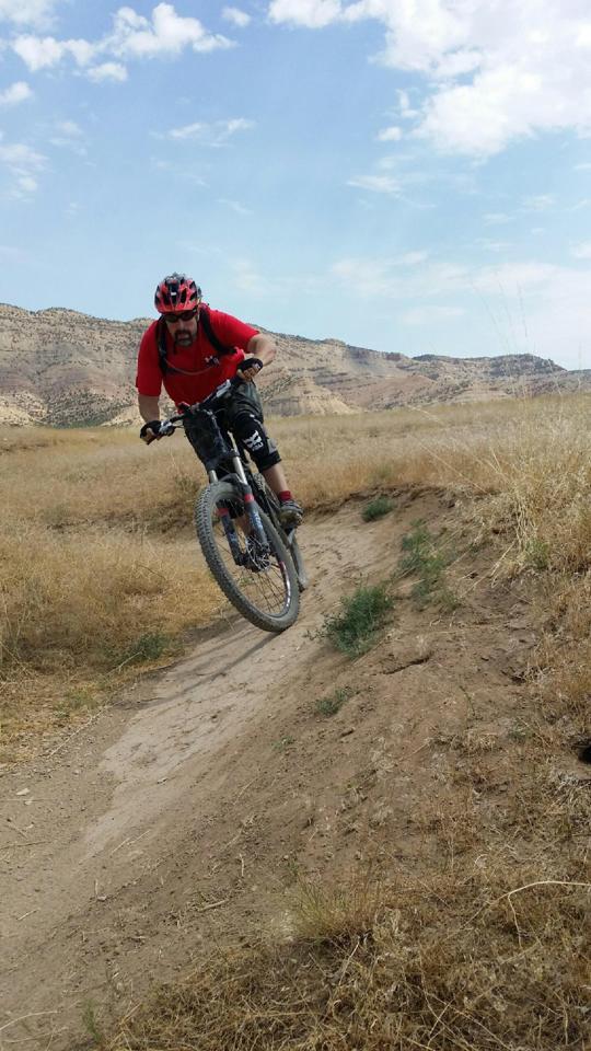 A mountain biker in a red shirt and helmet performs a jump on a dirt trail surrounded by dry grass and hills under a partly cloudy sky. Kessel Run mountain bike trail.