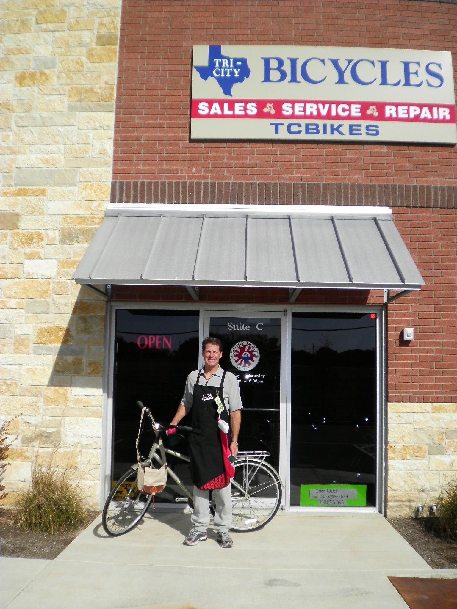 A man wearing a black apron stands outside a bicycle shop, holding a bicycle. The shop, named "Tri-City Bicycles," features a sign highlighting sales, service, and repair. The storefront displays an "OPEN" sign, and the building has a combination of brick and stone exterior. Nearby, there are shrubs and a clear sky in the background.