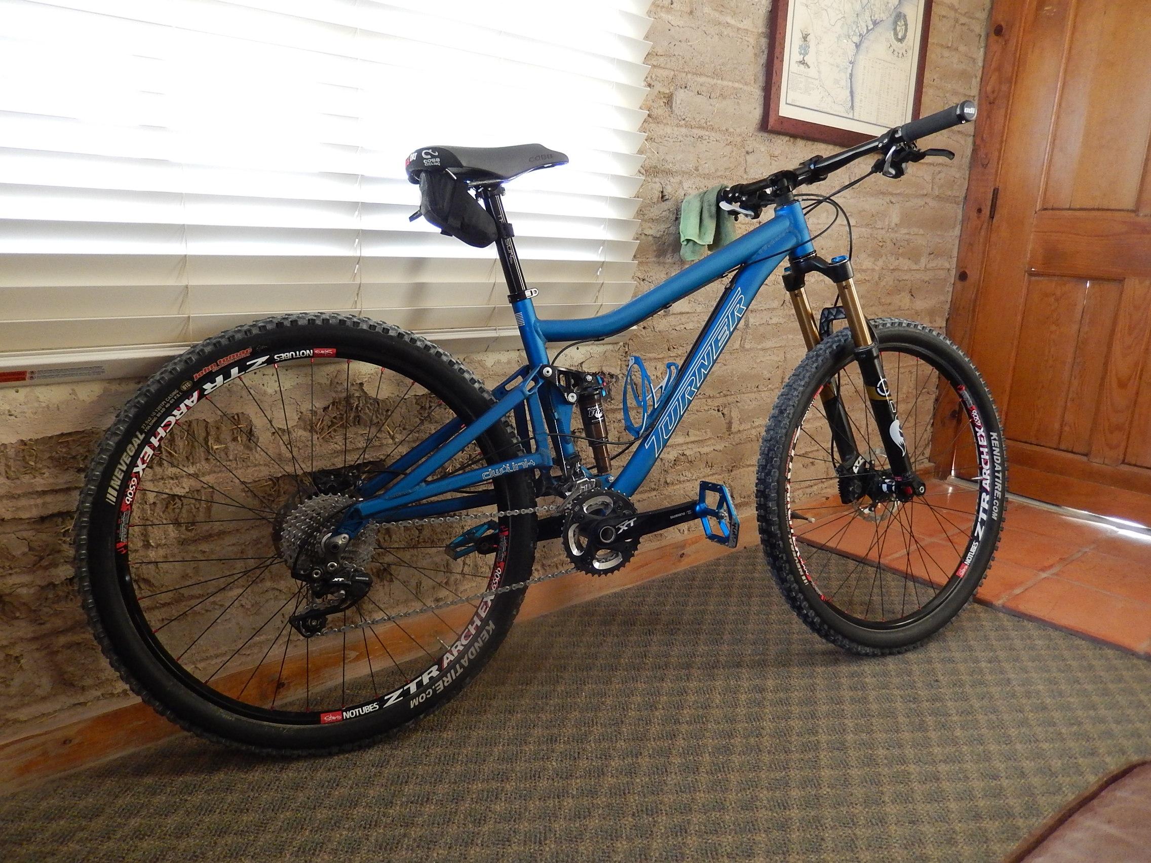 Turner Flux: A blue mountain bike leaning against a brick wall, featuring a black seat and black handlebars. The bike has thick, knobby tires and a visible gear setup, with a wooden door and light-colored blinds in the background.
